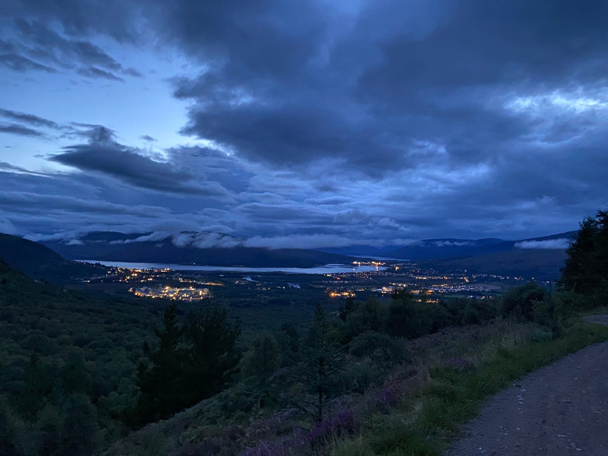 Fort William and Loch Linnhe lit up at dusk under dark blue clouds, seen from the forest track