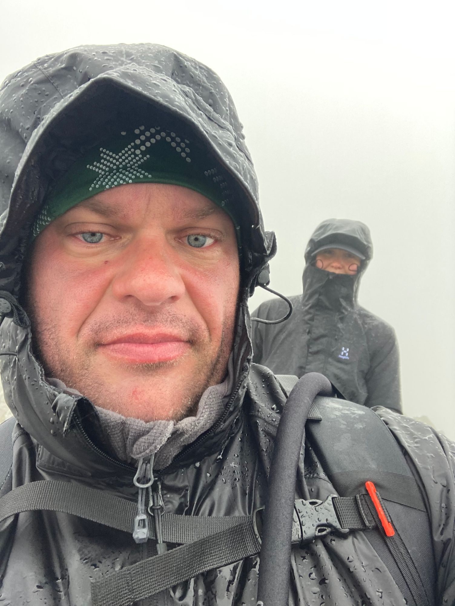 Close-up selfie of Leif soaked in a rain hood, with Kristine behind him masked against the wind and rain