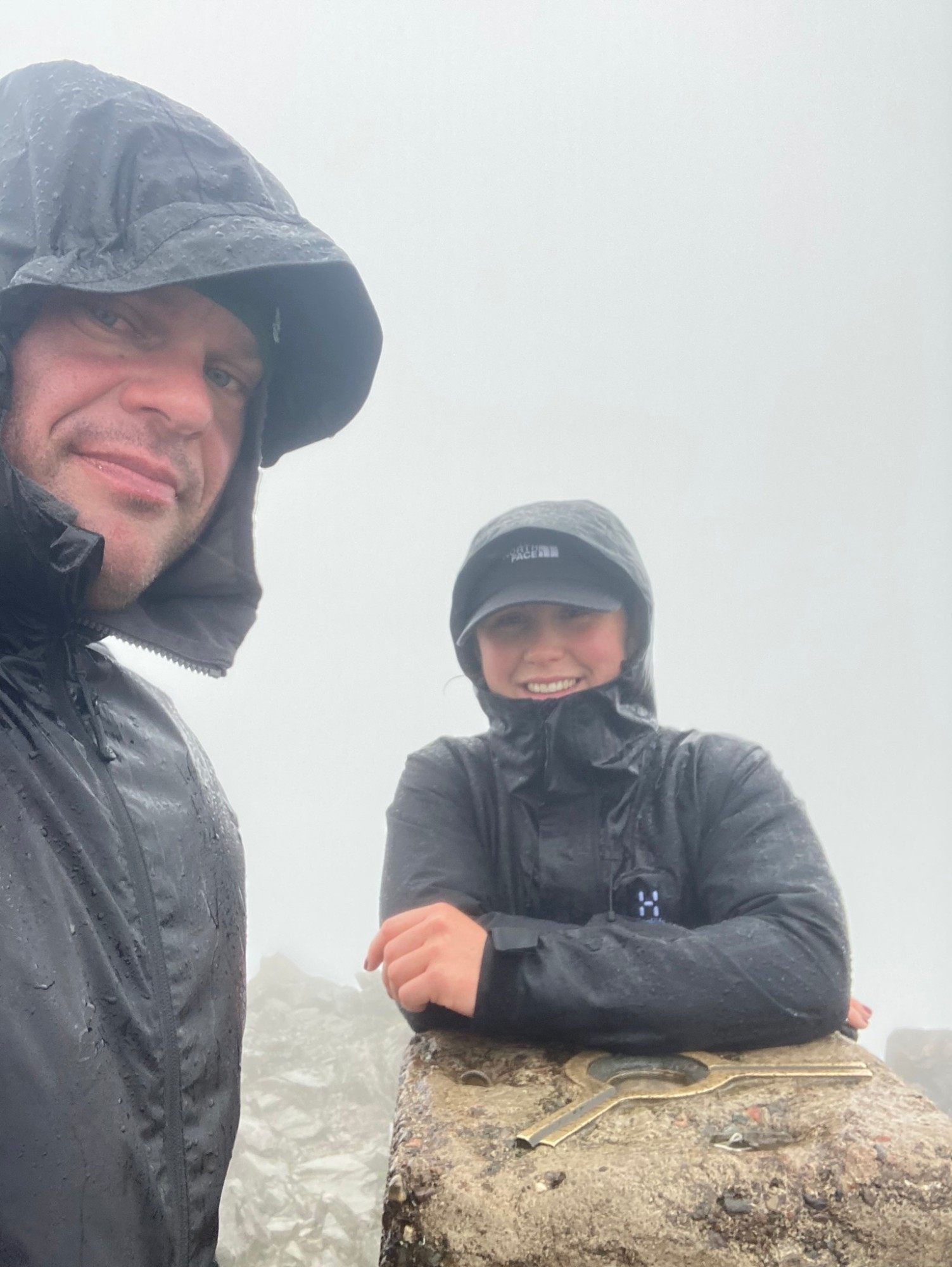 Leif and a smiling Kristine leaning on the trig pillar in rain and cloud