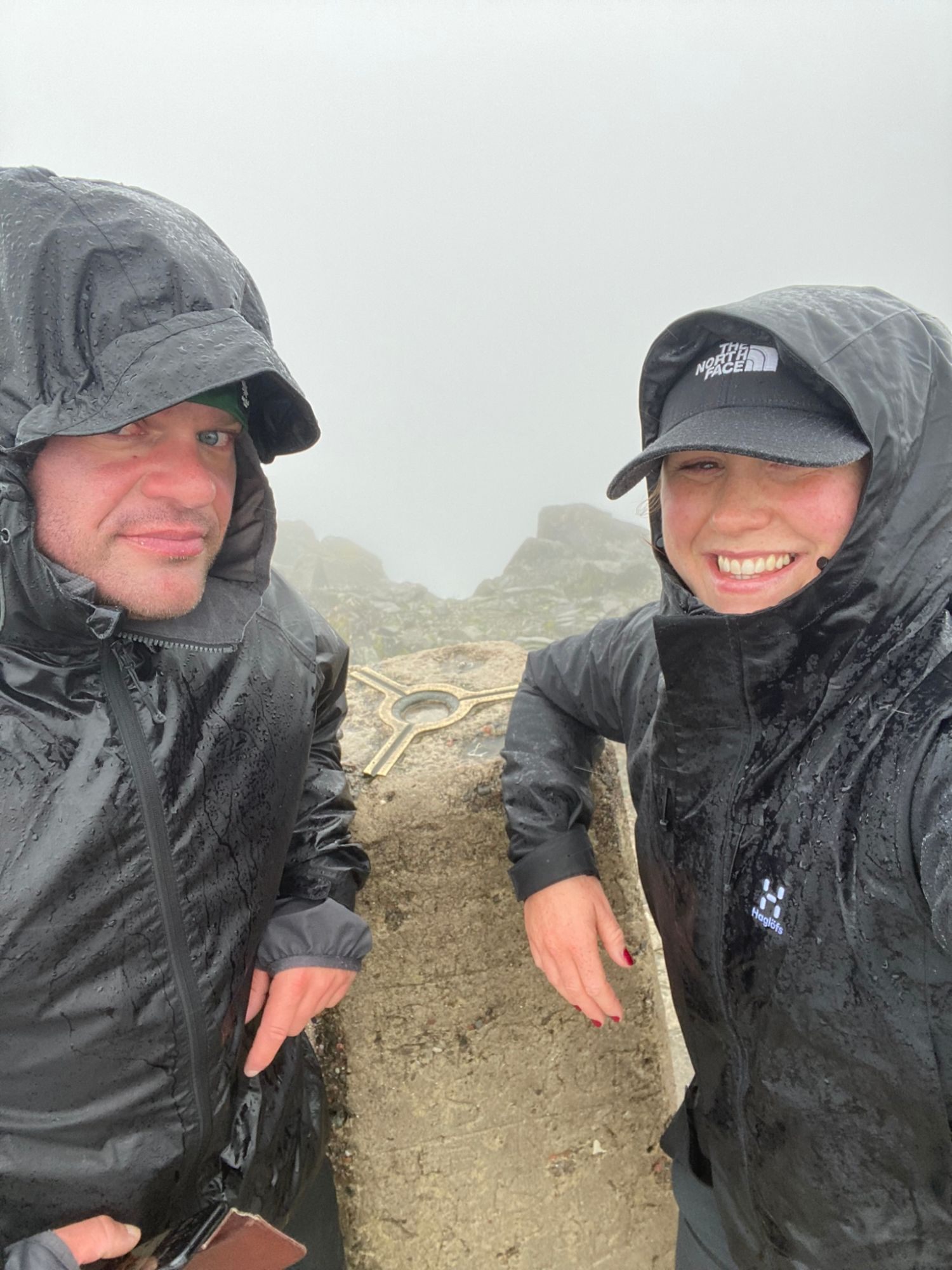 Leif and Kristine soaked in rain hoods on either side of the Ben Nevis trig pillar, Kristine grinning
