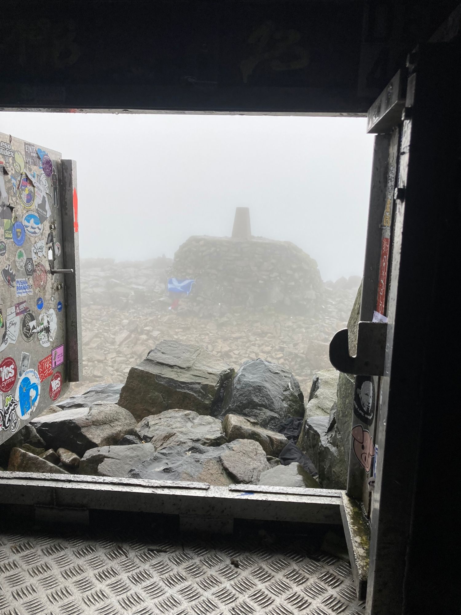 View out of the summit shelter door, covered in stickers, towards the trig point cairn in fog with a Scottish flag