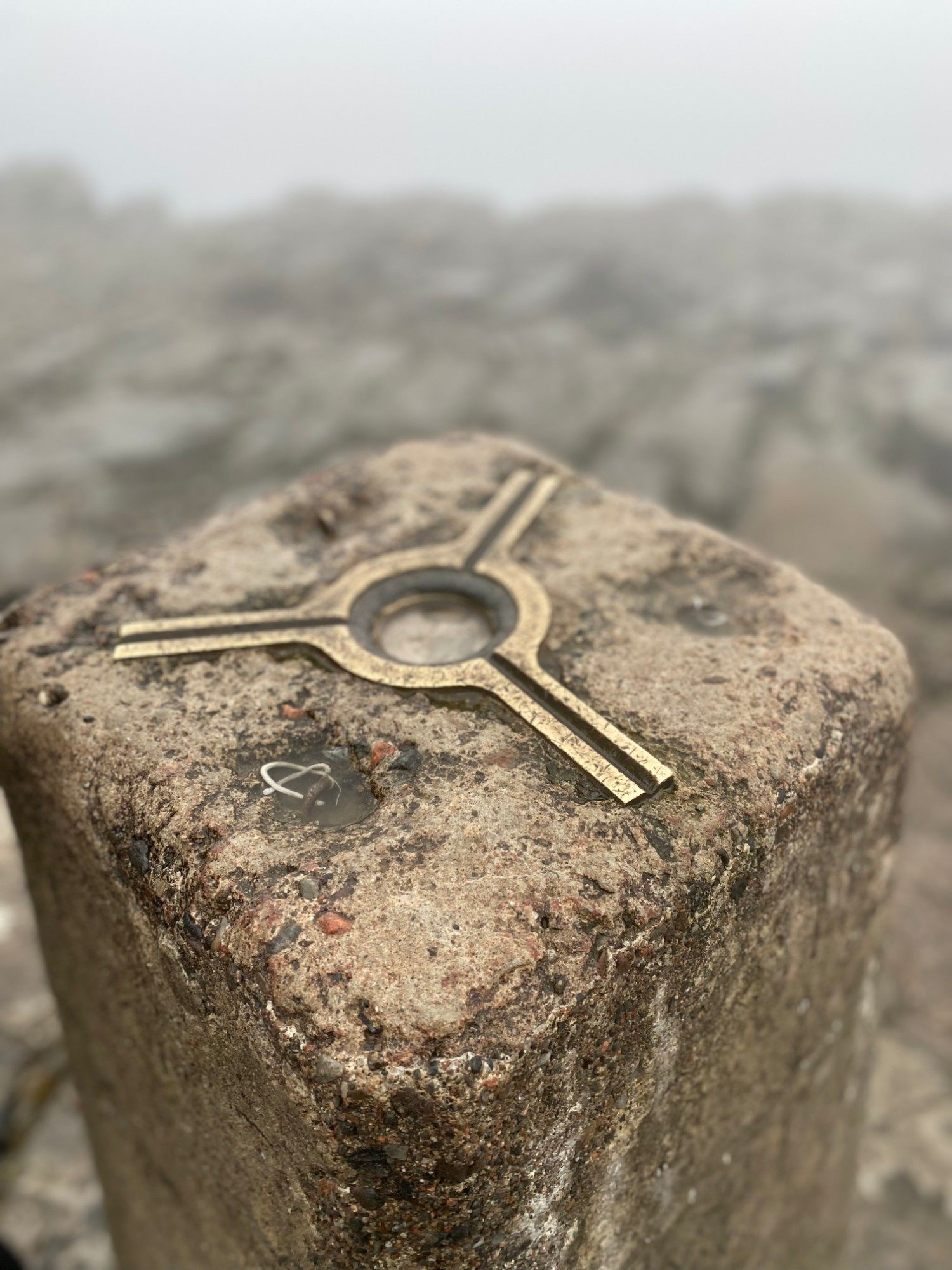 Close-up of the brass plate on top of the Ben Nevis trig pillar, with rocks fading into fog