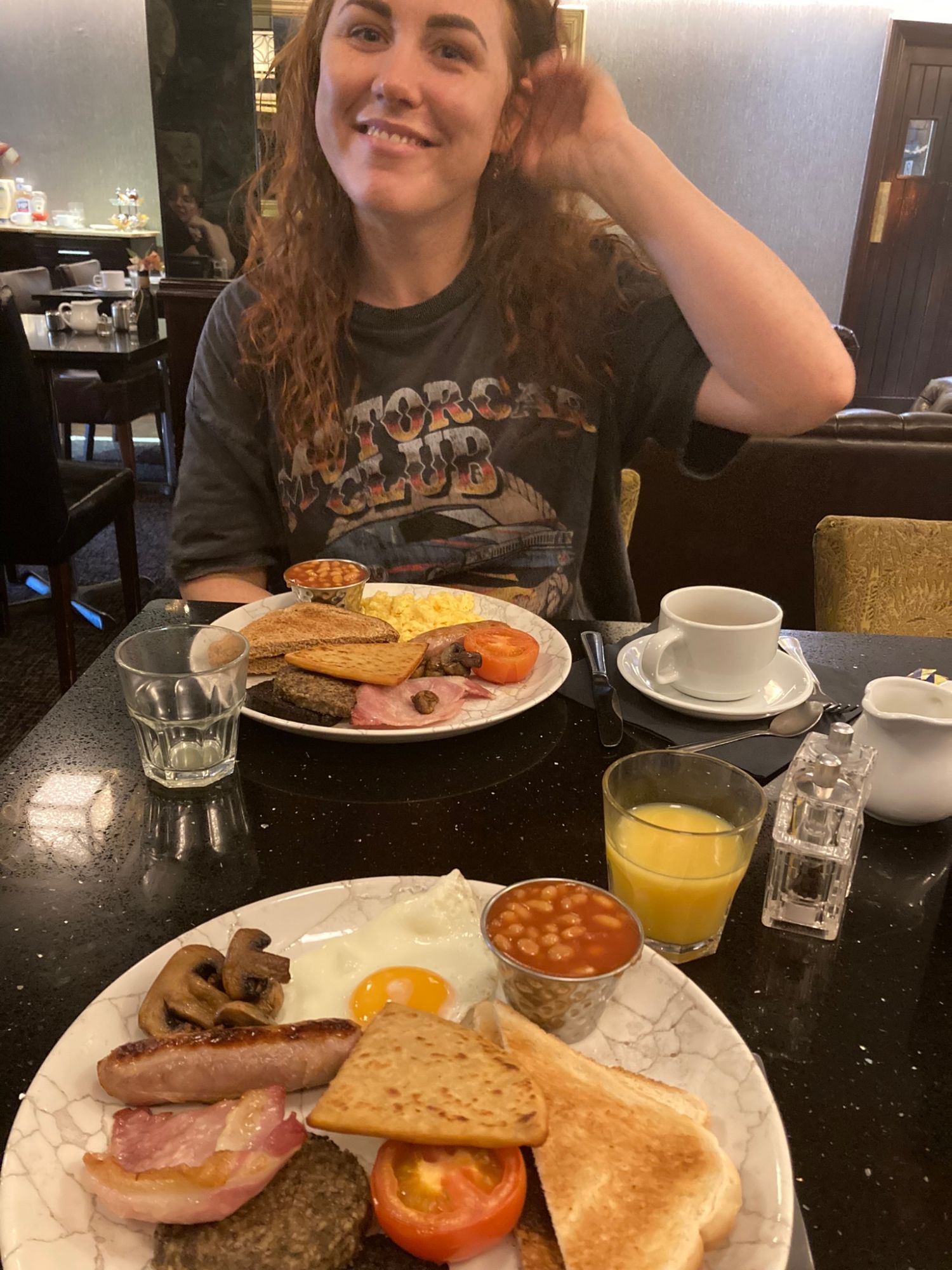 Kristine smiling at the breakfast table behind a full cooked breakfast with egg, bacon, sausage, beans, tomato, mushrooms and toast
