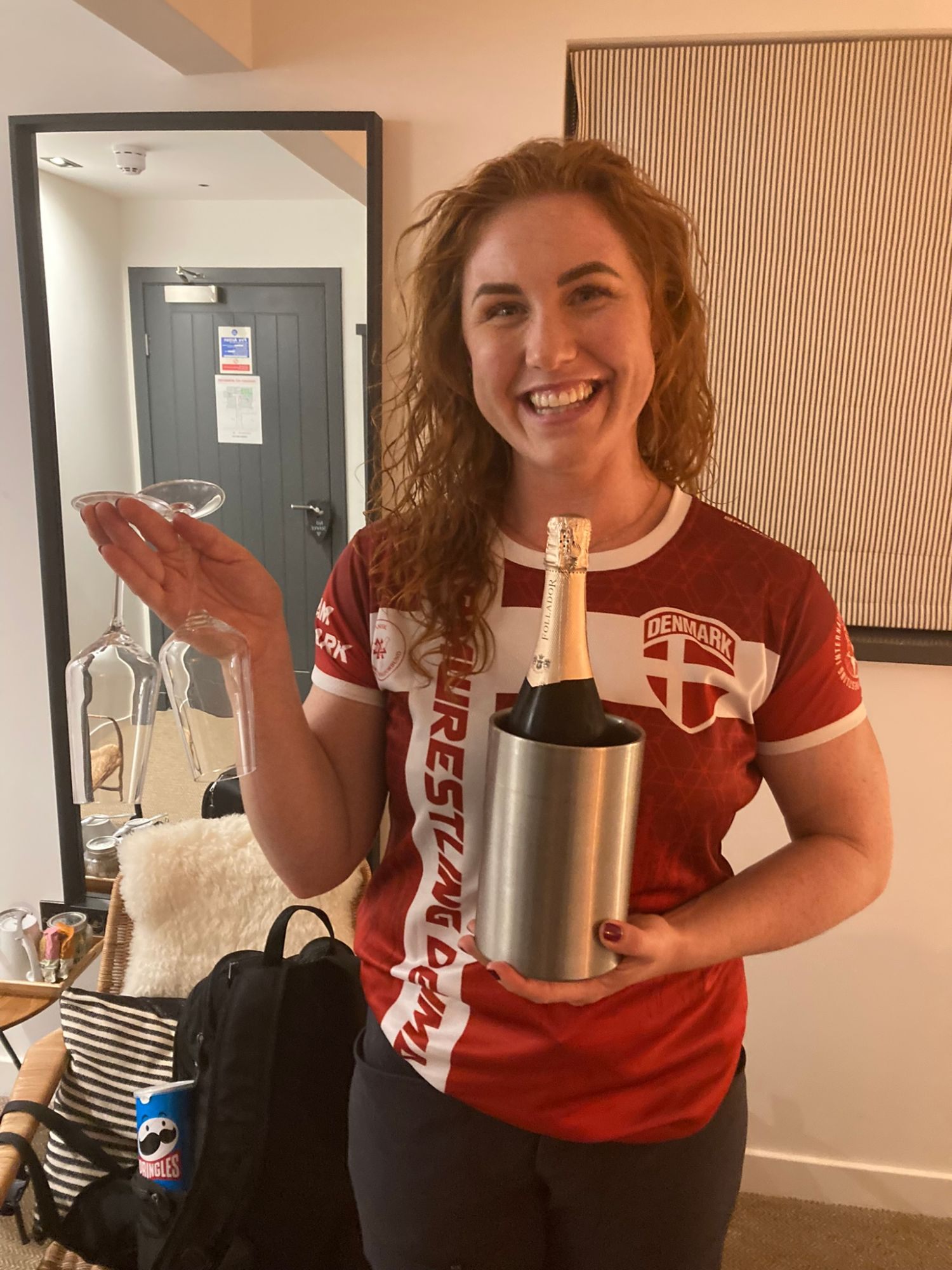Kristine smiling in a Danish arm wrestling shirt, holding a bottle of bubbly in a cooler and two glasses