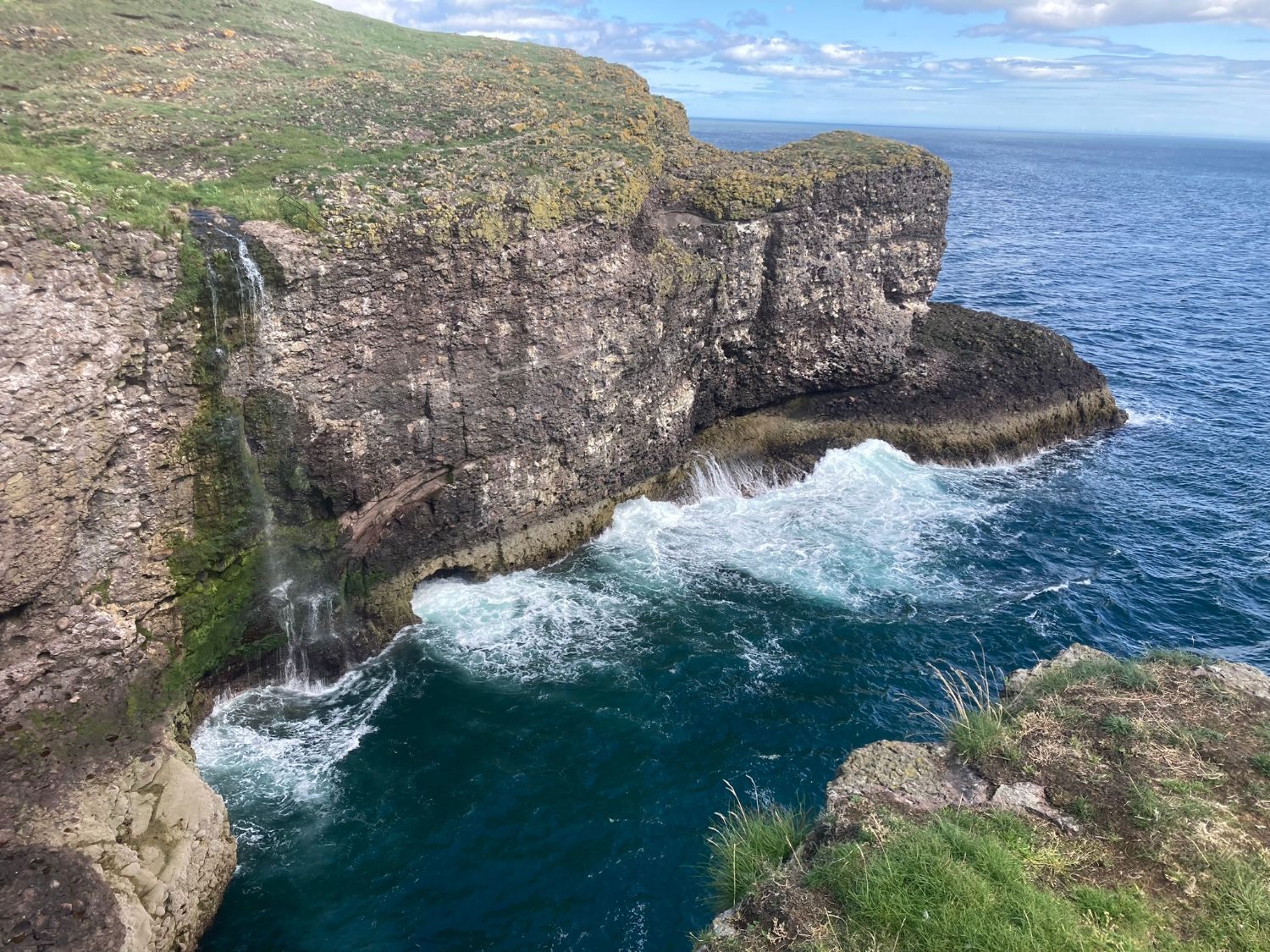 Crawton Waterfall falling from a grassy clifftop into a deep blue cove with white surf, Aberdeenshire