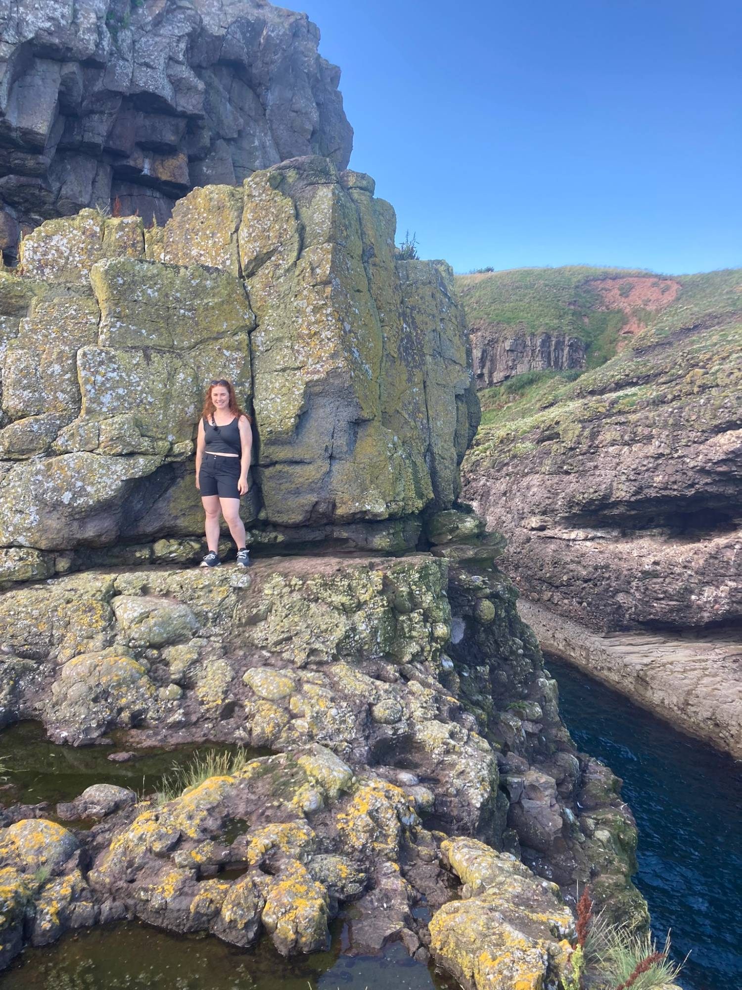 Kristine standing on a ledge of lichen-covered rock above a pool and the sea