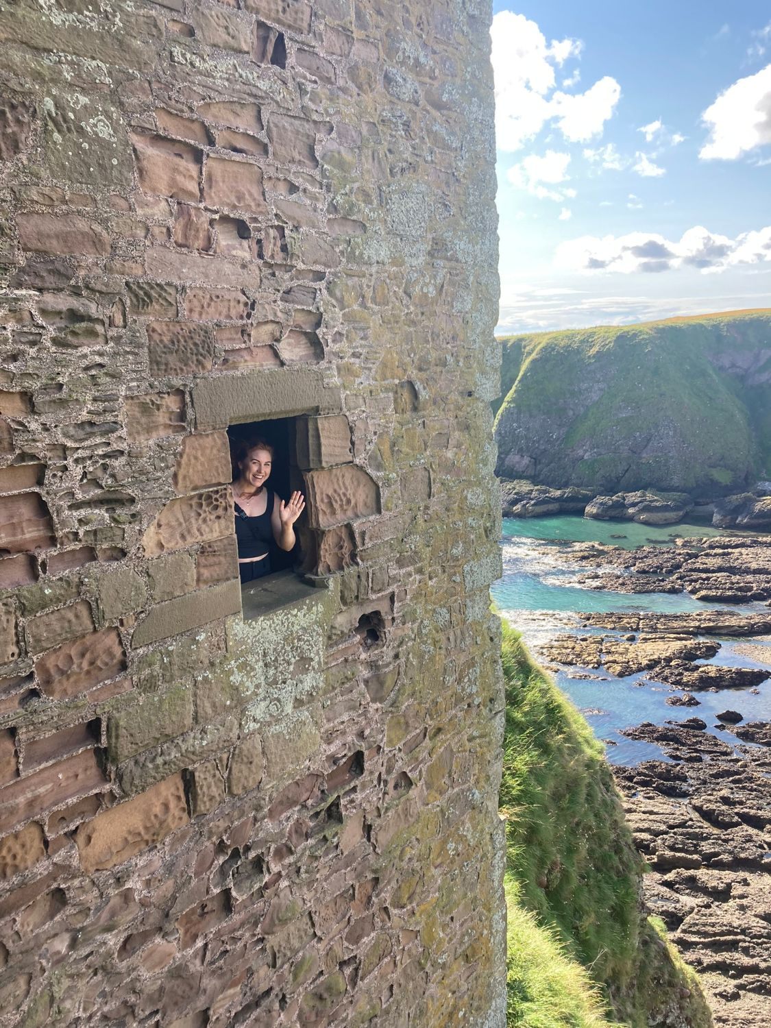 Kristine waving from a small window in a weathered sandstone wall, with turquoise sea and rocks below