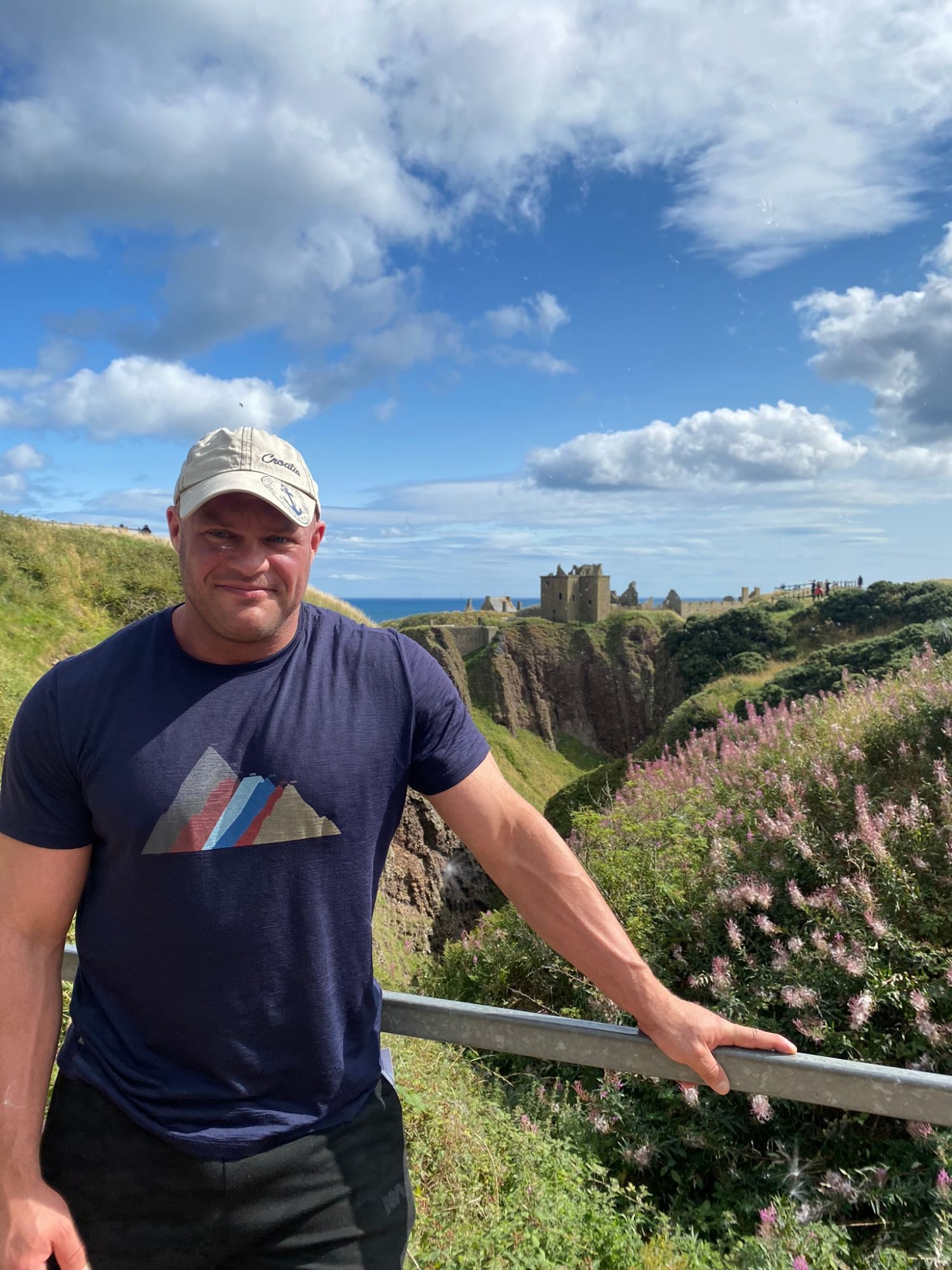 Leaning on a railing above the gorge with Dunnottar Castle and the sea behind, pink flowers on the slope