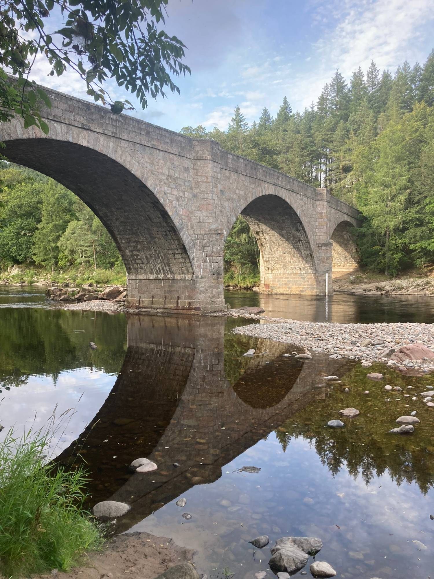 The stone arches of Potarch Bridge over the River Dee, reflected in calm water with trees behind