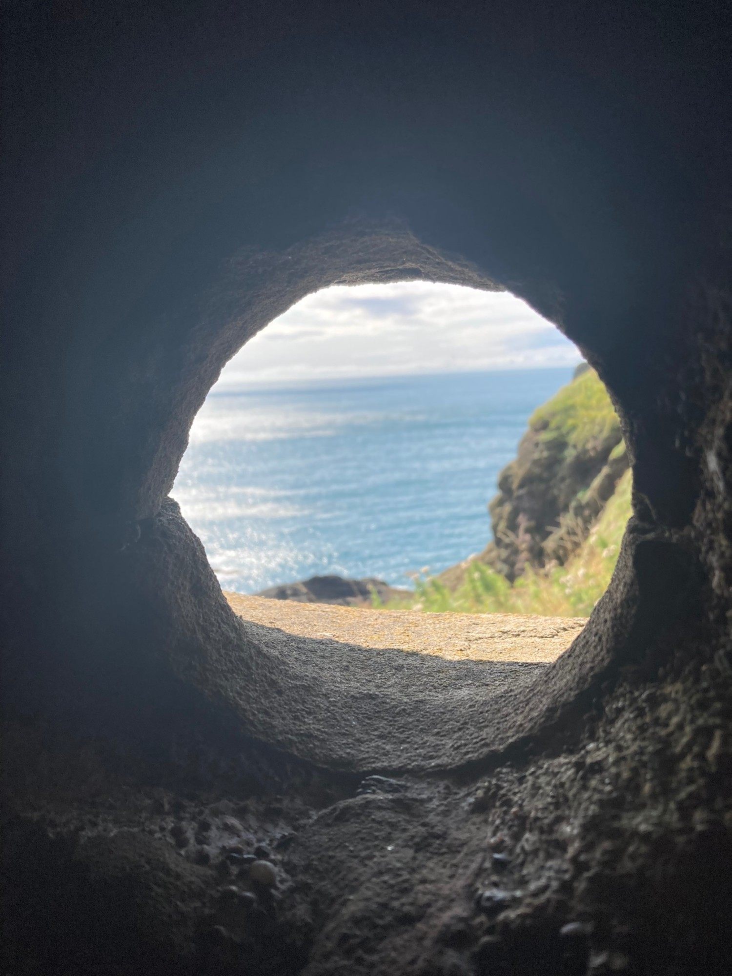 The sea and a green cliff seen through a round opening in a thick stone wall