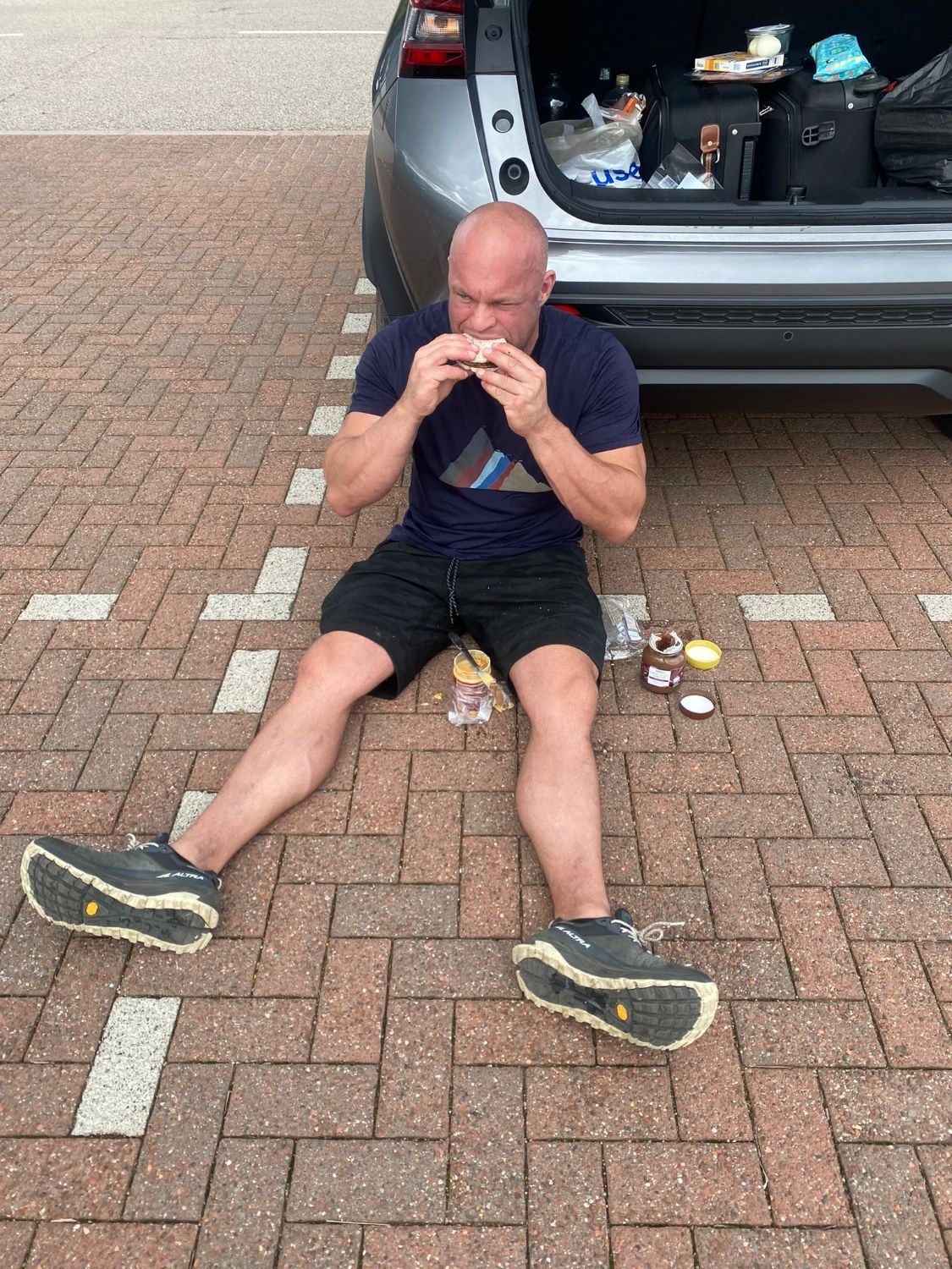 Sitting on the paving behind the open boot of the rental car, eating a rice cake with jars of spread beside me