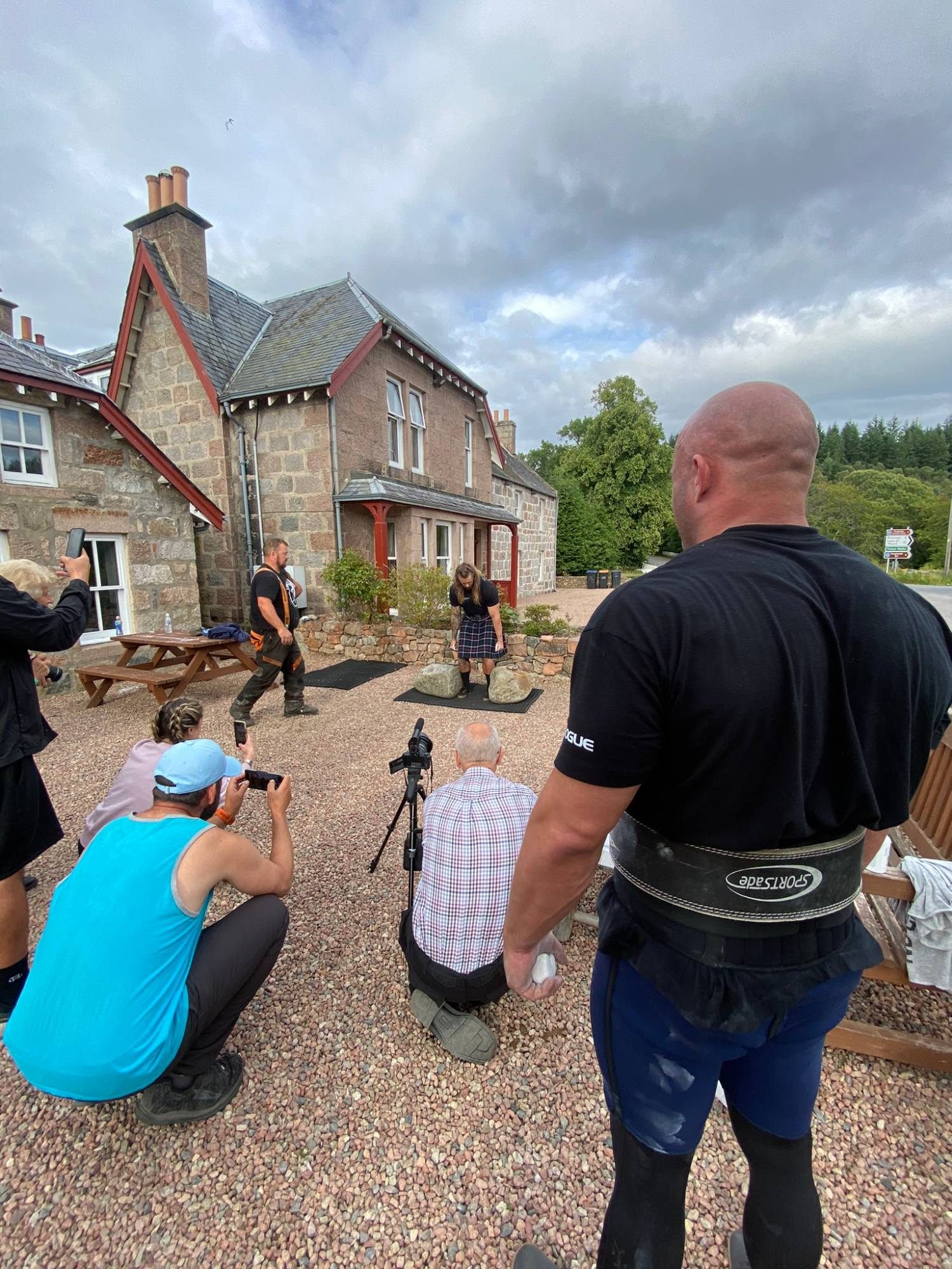 Waiting with chalk in hand, wearing a lifting belt, while another lifter in a kilt sets up over the stones and people film from the gravel
