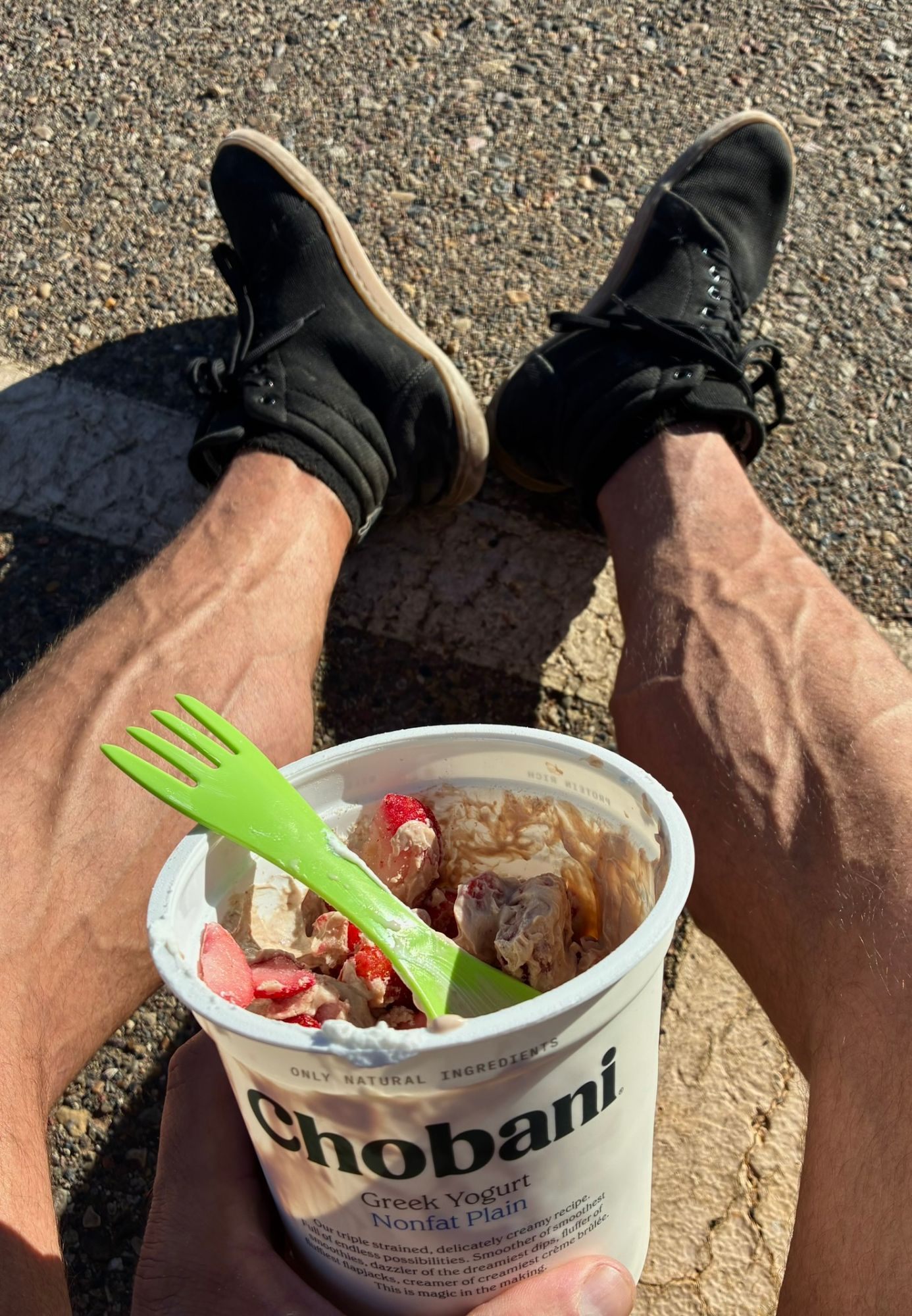 A tub of plain Greek yoghurt with strawberries and a green plastic spork, held over legs in black trainers on a sunny car park