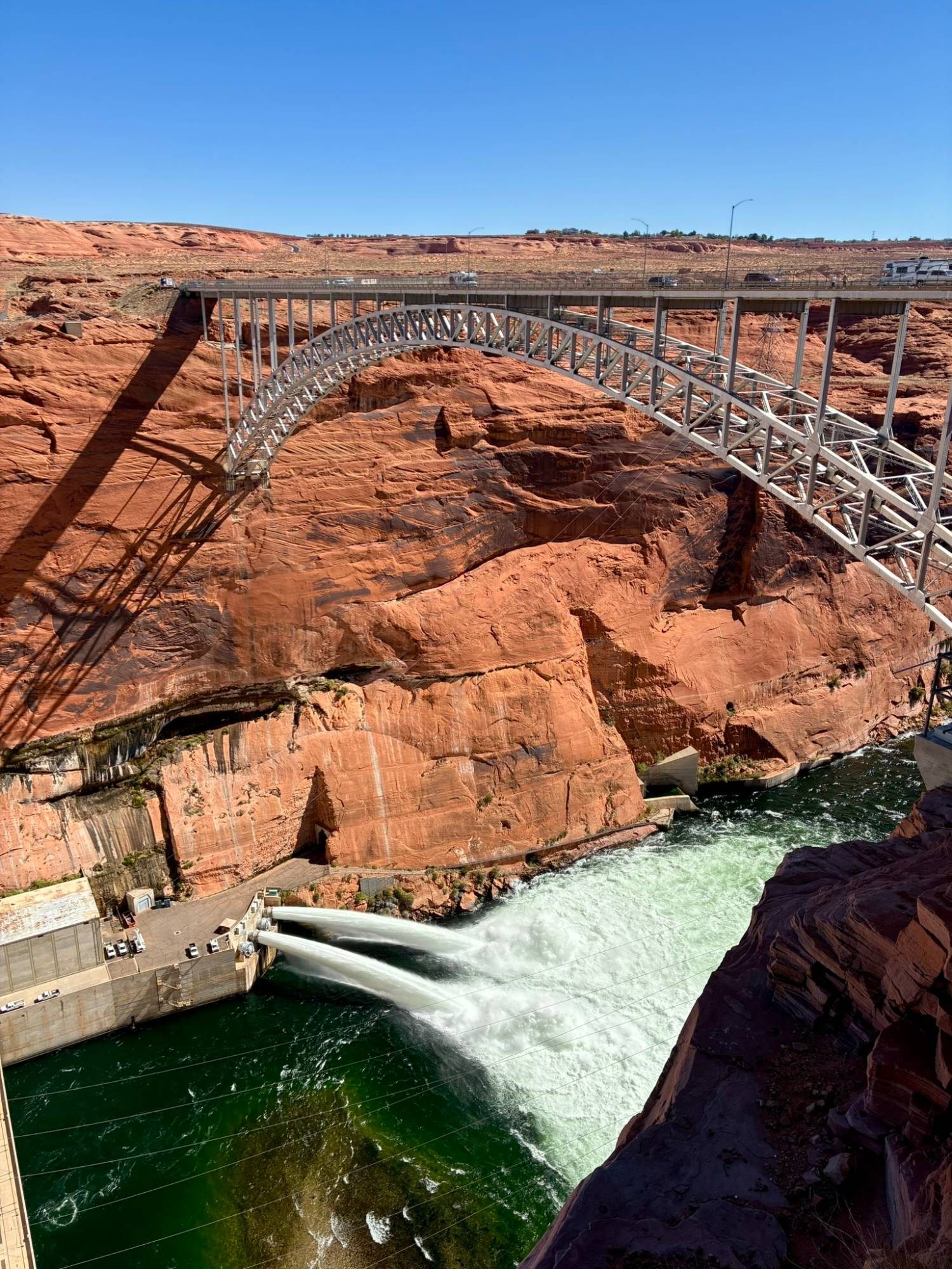 The steel arch of Glen Canyon Dam Bridge over red sandstone, with two jets of water shooting into the green Colorado River below the dam