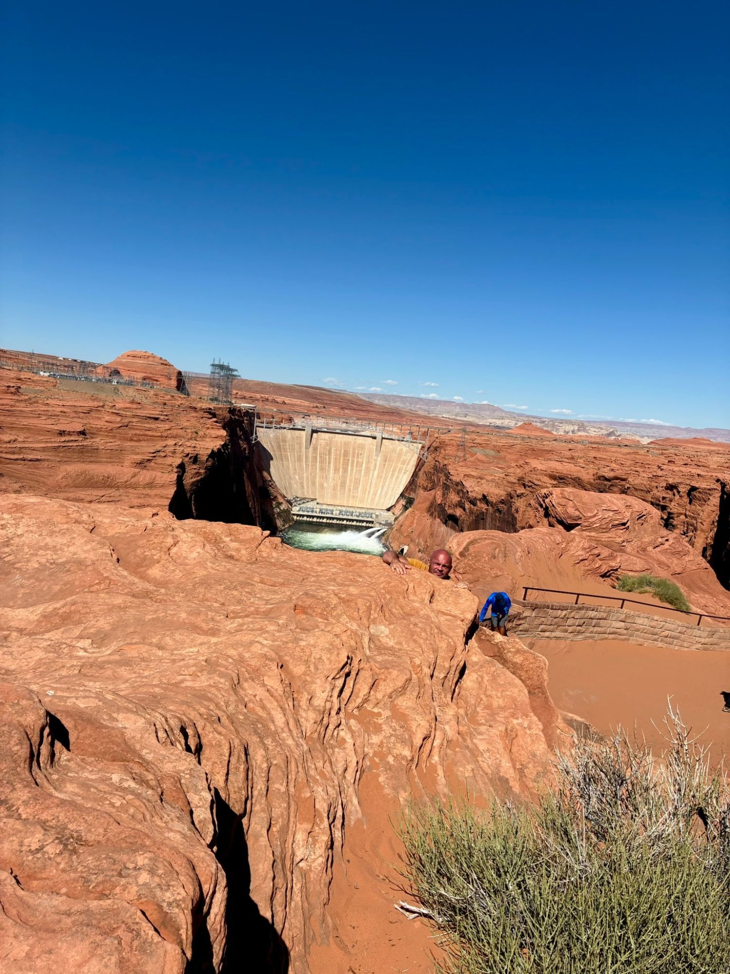 Leif peeking up from behind a sandstone boulder, with Glen Canyon Dam and its bridge behind