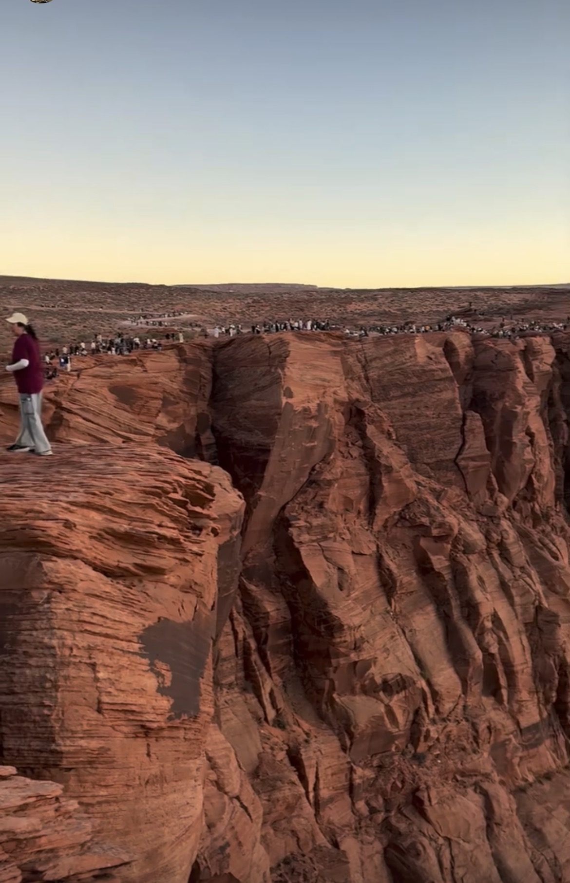 A long line of visitors crowded along the rim of the main Horseshoe Bend viewpoint at sunset, seen from further along the canyon