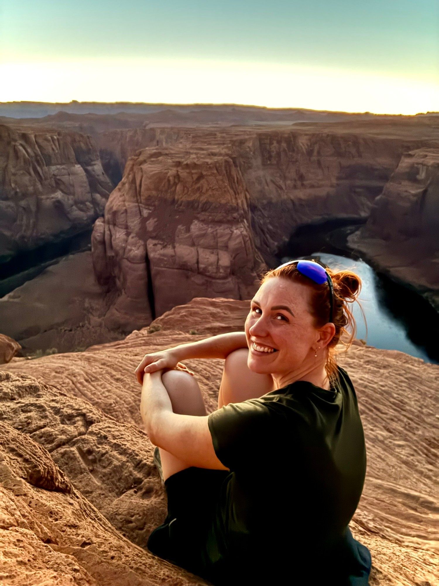 Kristine smiling over her shoulder, sitting on the rim with Horseshoe Bend behind her