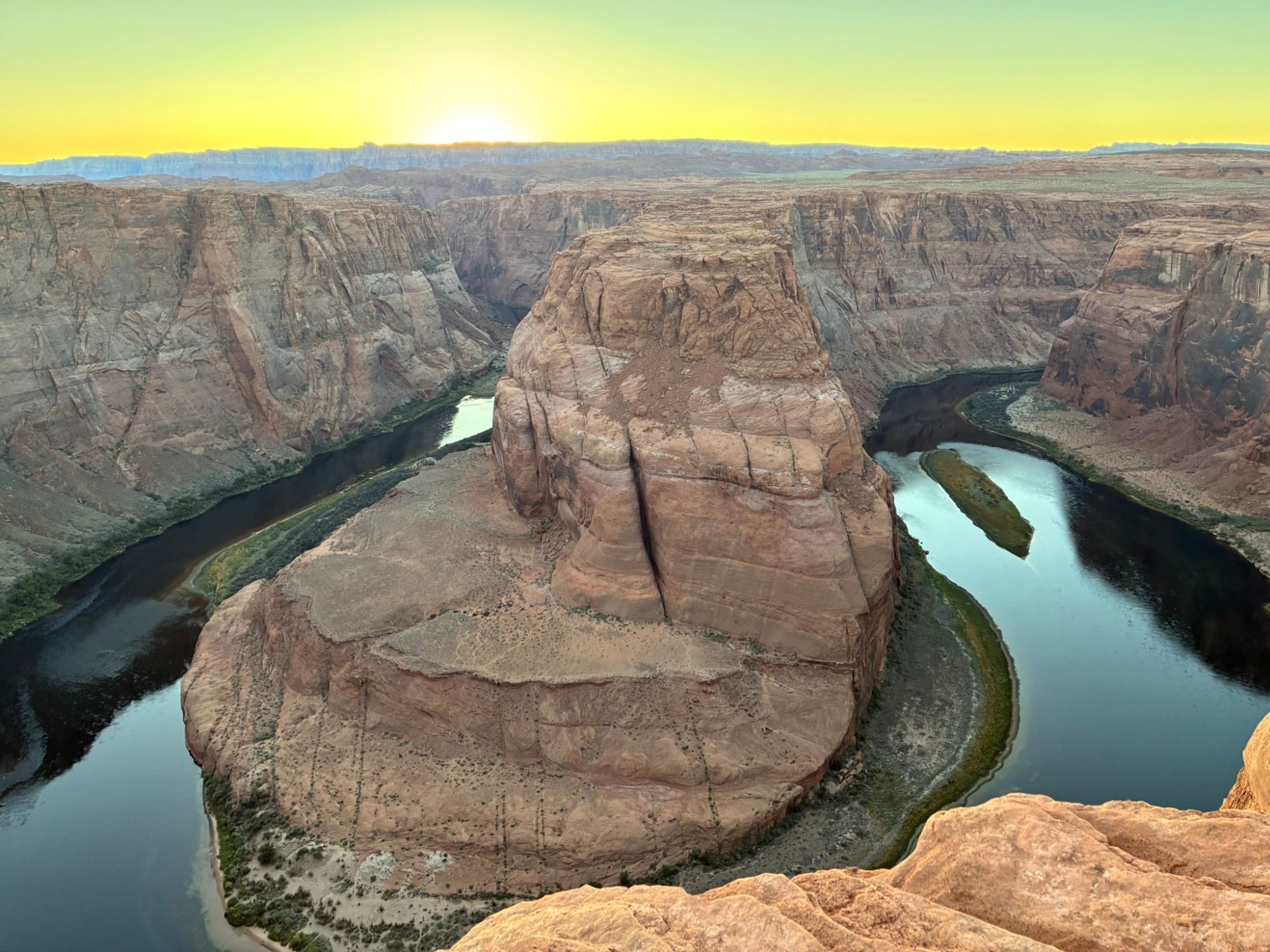 The sun setting on the horizon behind Horseshoe Bend, with the river reflecting the sky