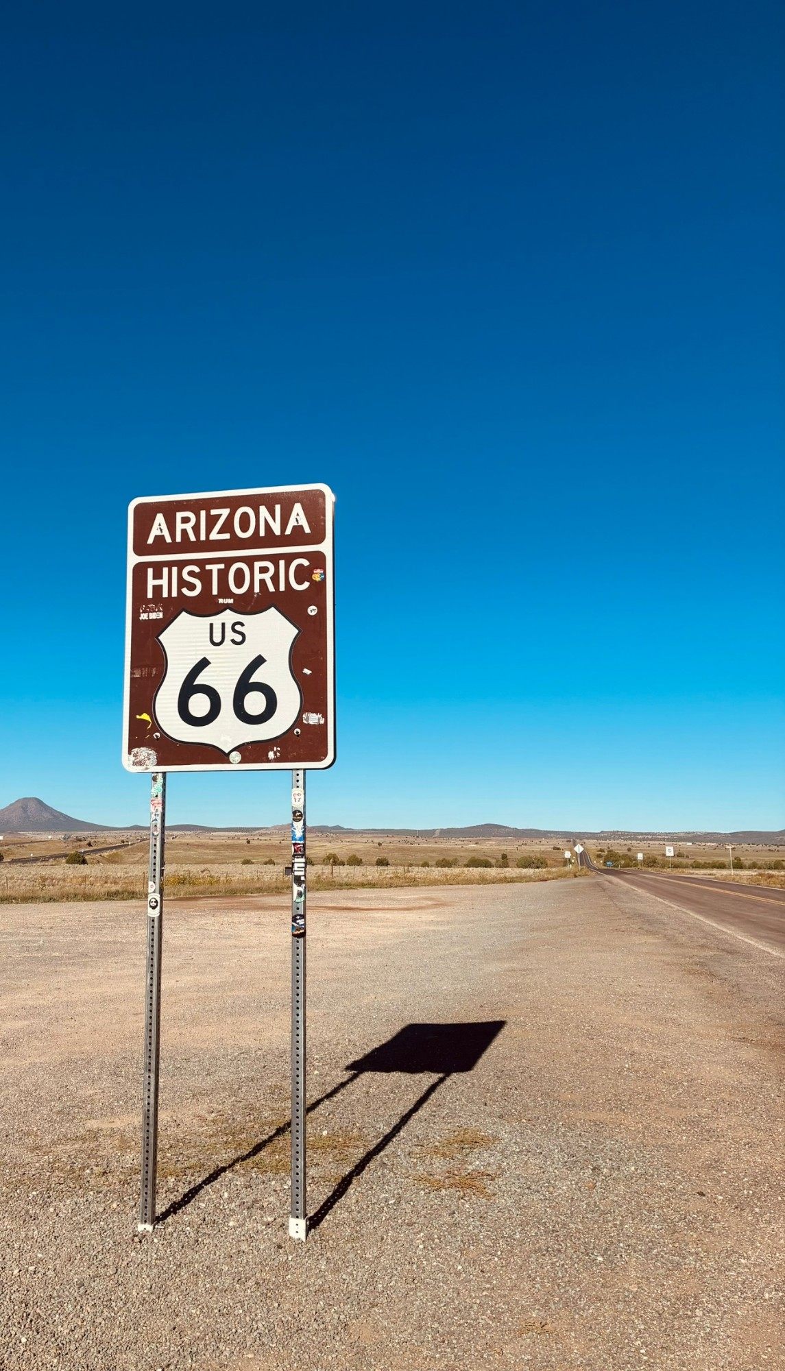 A brown Arizona Historic US 66 sign on a gravel verge beside an empty road stretching into the desert under a deep blue sky