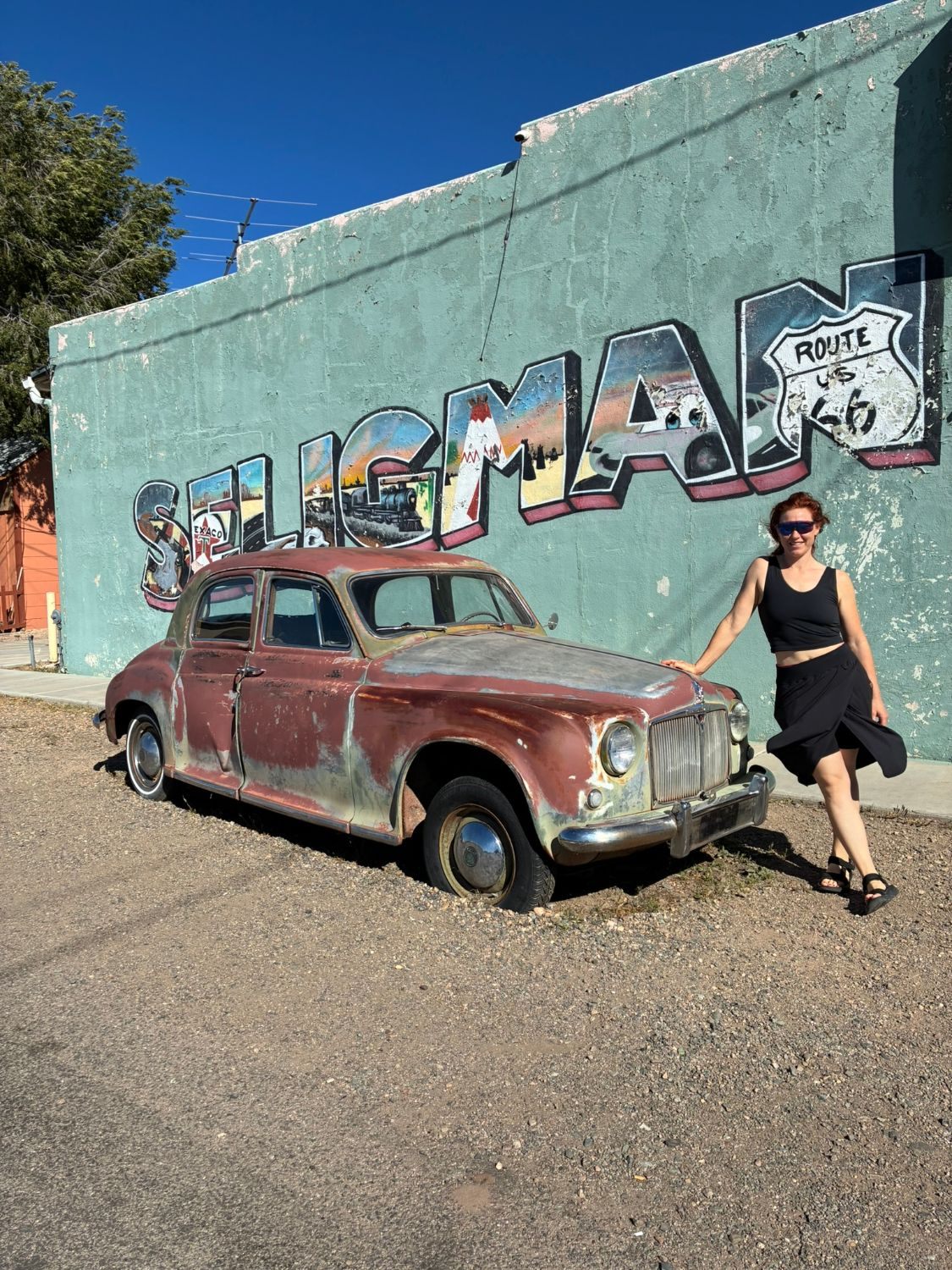 Kristine standing beside a rusty vintage car in front of a big SELIGMAN mural with a Route 66 shield