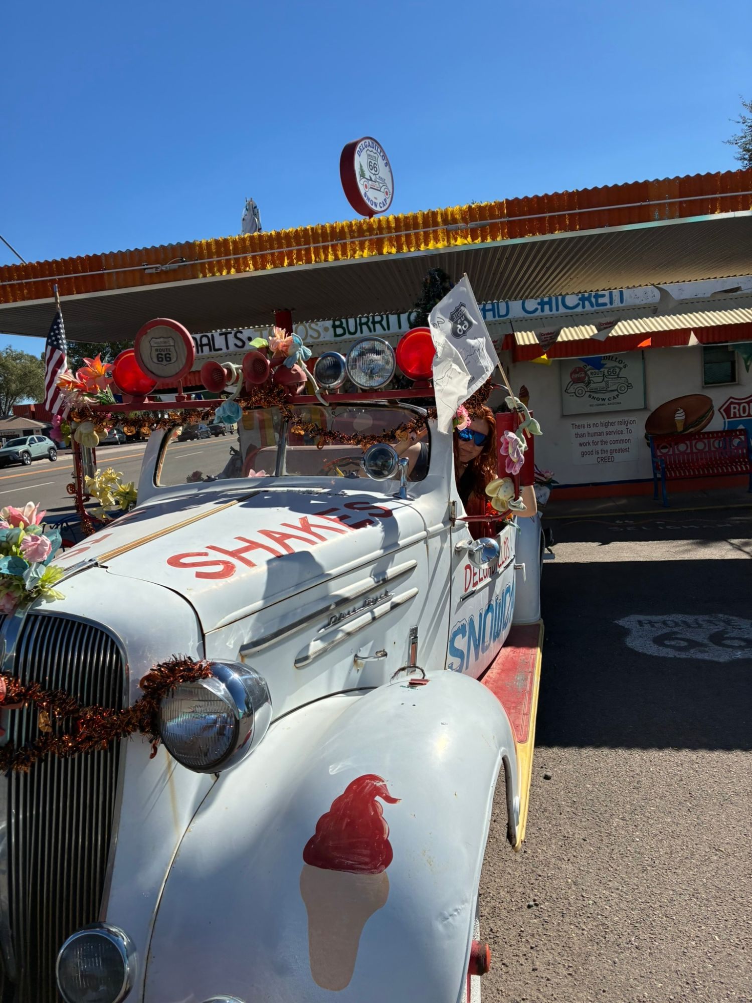 Kristine waving a Route 66 flag from the driver's seat of the decorated white Snow Cap car covered in flowers