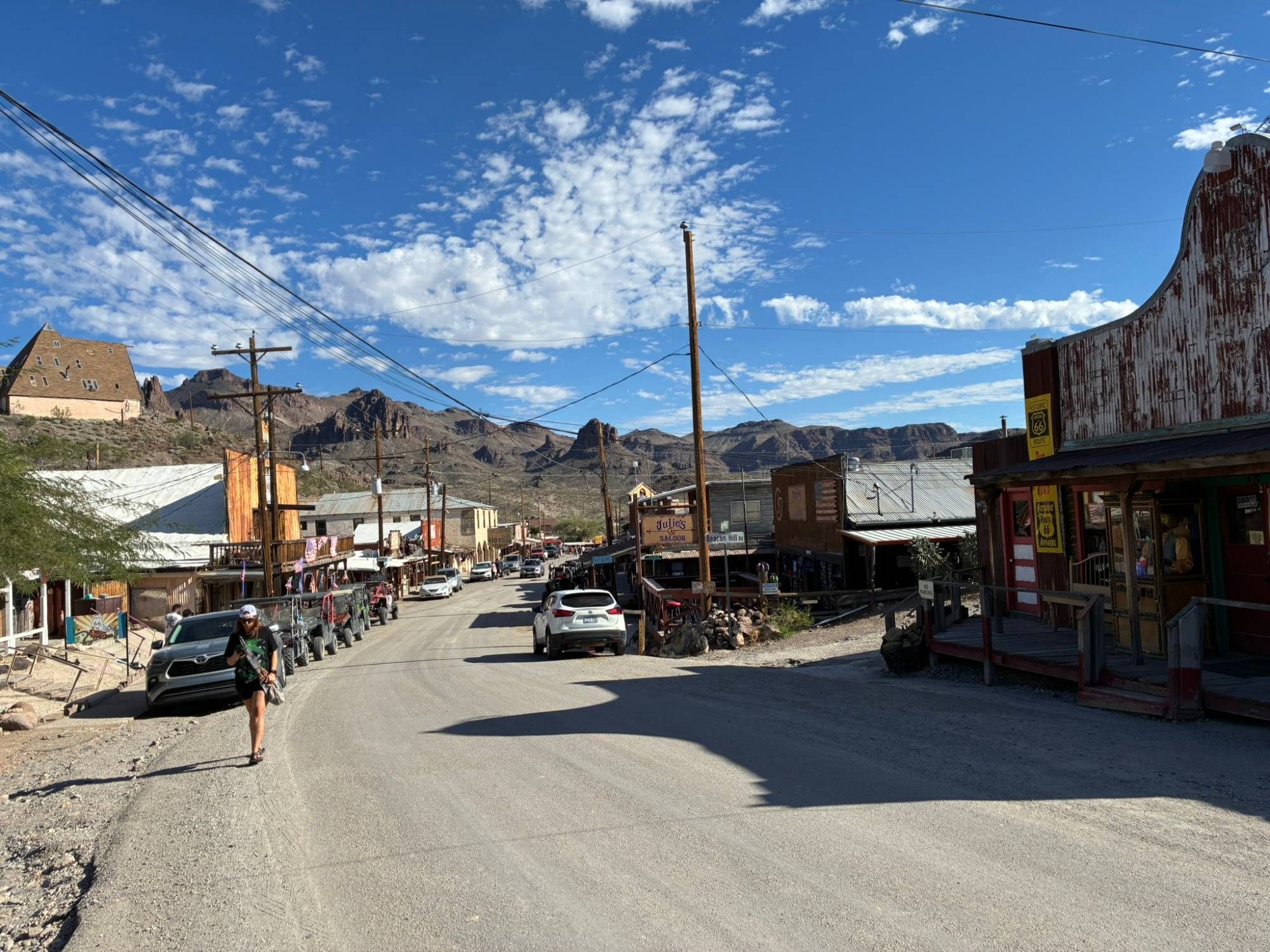 The dusty main street of Oatman with wooden western buildings, Julie's Saloon sign and desert mountains behind, Kristine walking in the foreground