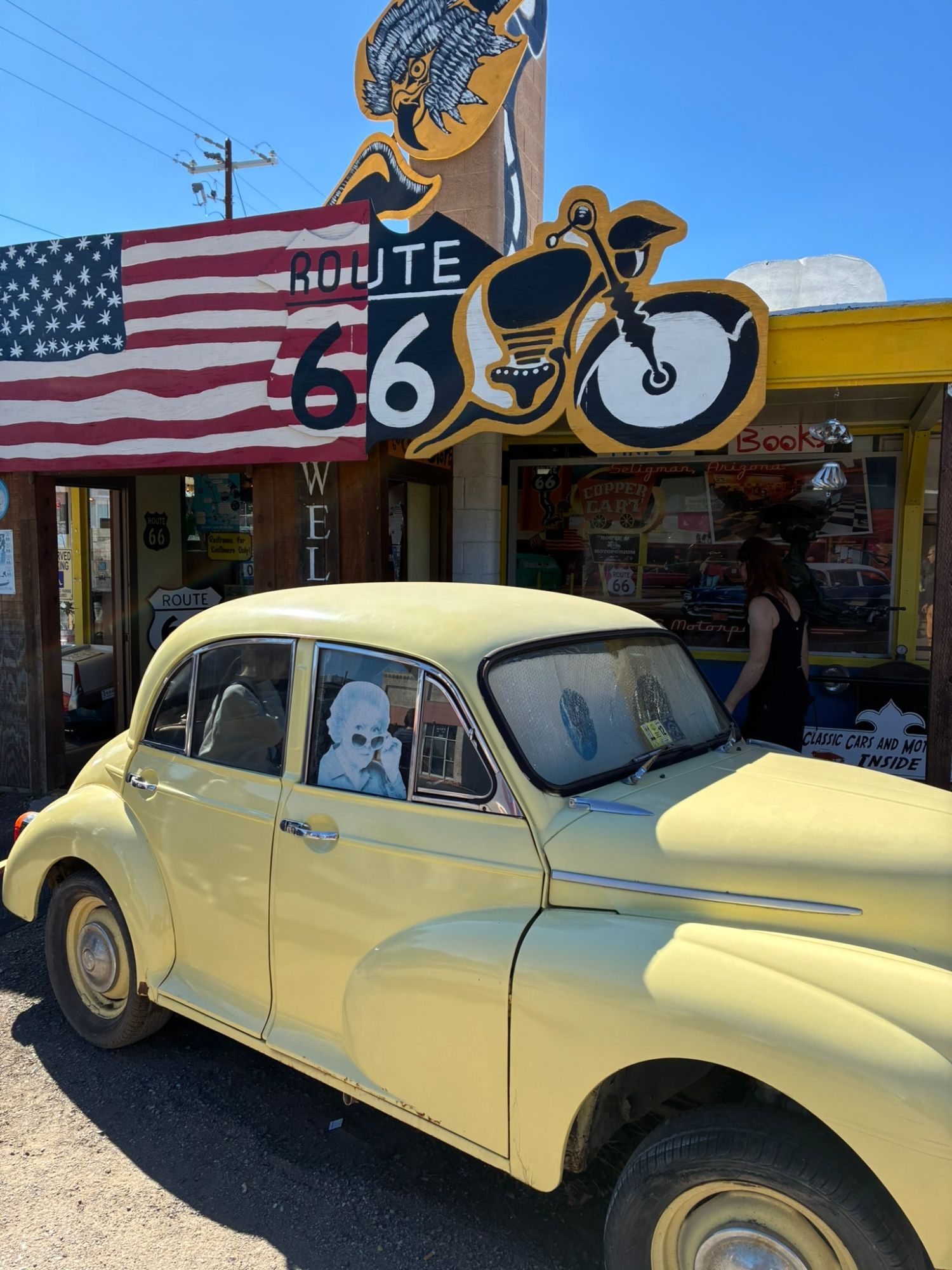 A pale yellow vintage car parked in front of a Route 66 shop with a painted American flag and a motorbike sign