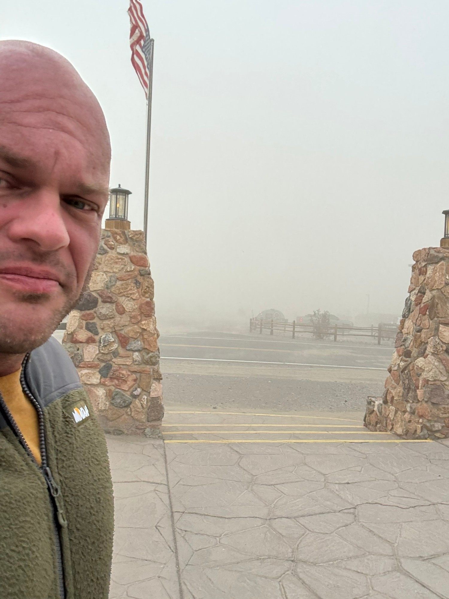 Selfie by the stone gateway at Stovepipe Wells, with the US flag and the road behind lost in blowing sand