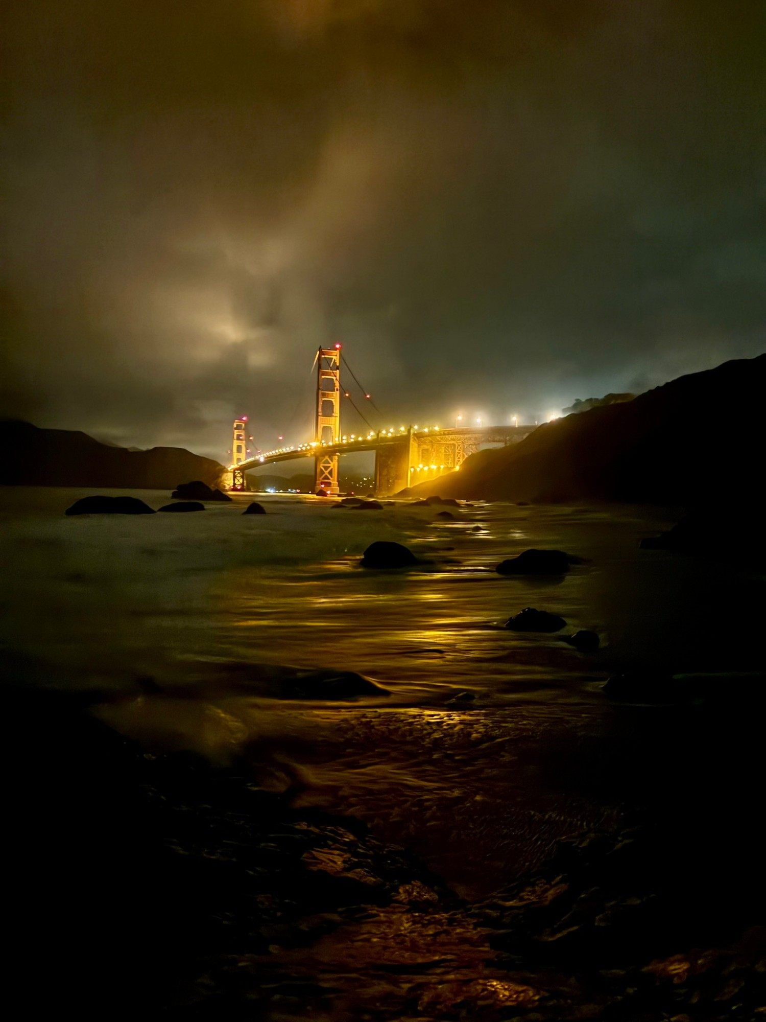 Waves washing over the rocks of Marshall's Beach with the lit-up Golden Gate Bridge reflected in the water