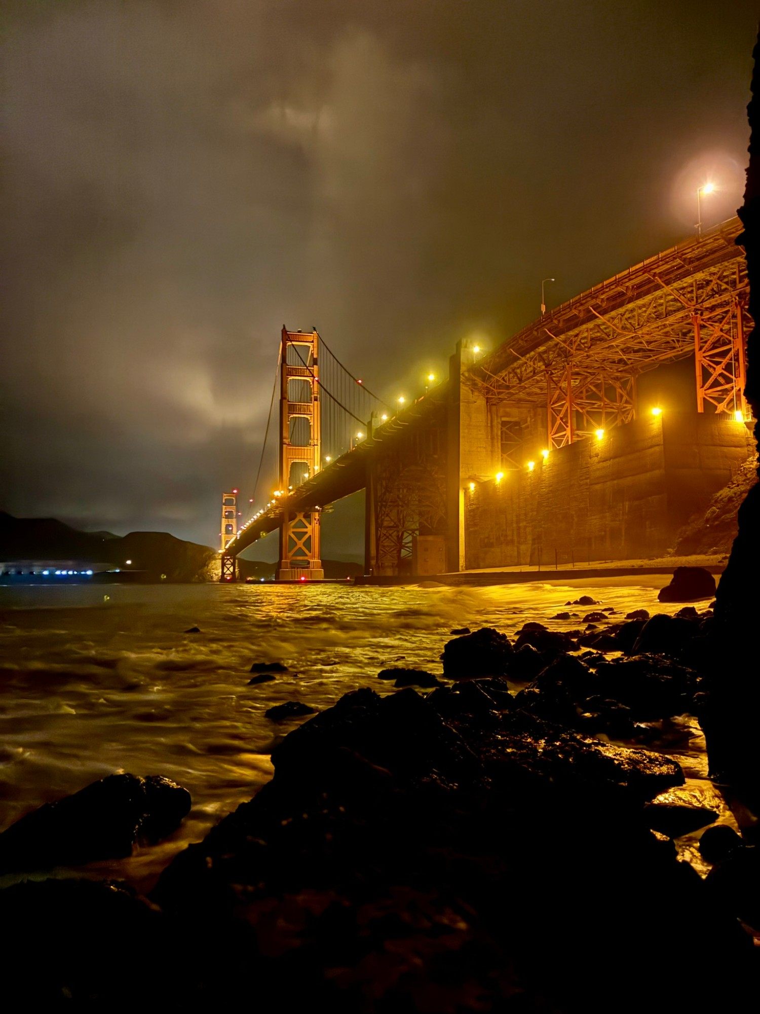 Close to the south end of the Golden Gate Bridge at night, with the lit-up steel structure above the waves and rocks
