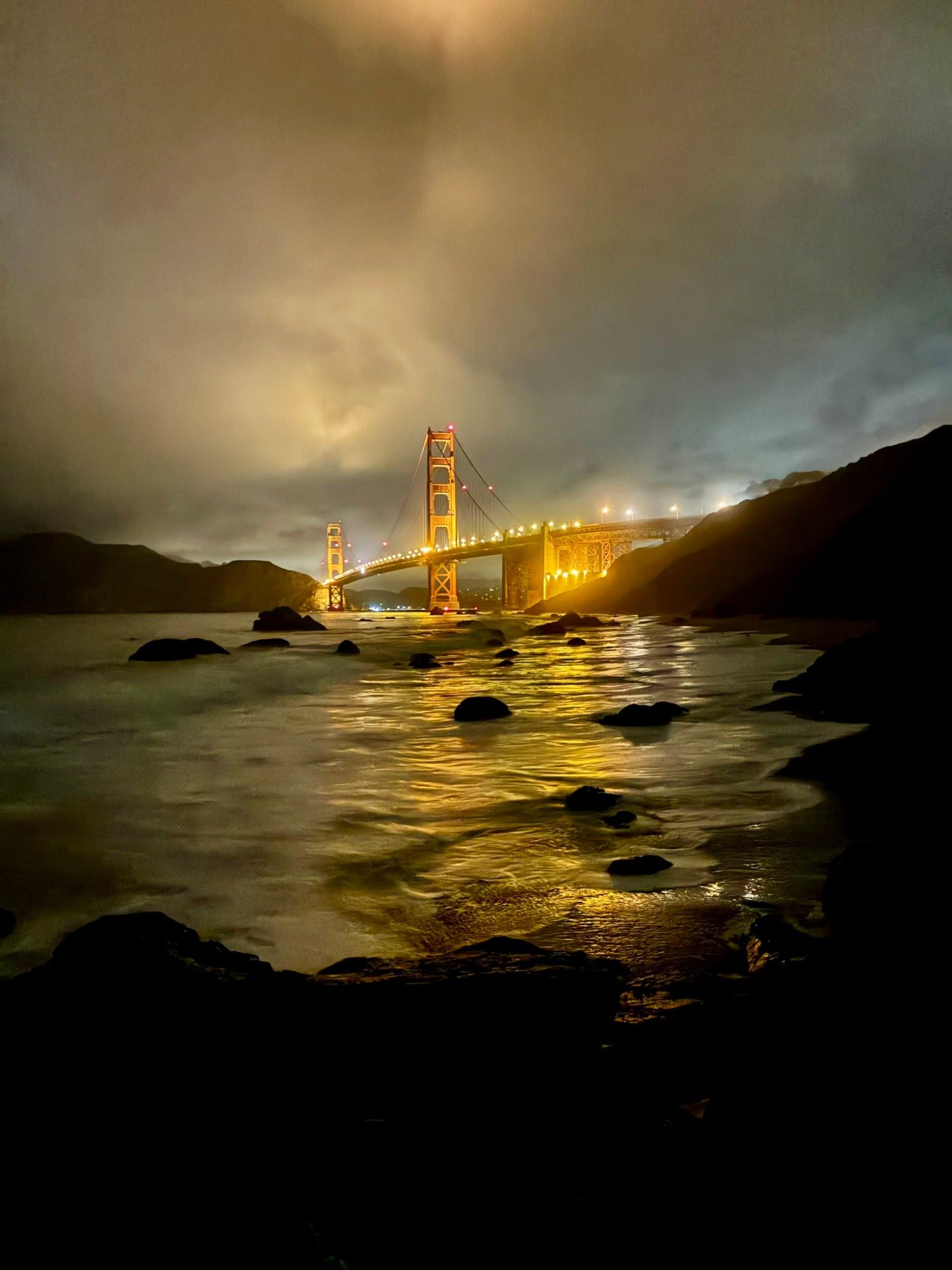 The Golden Gate Bridge lit up at night seen from Marshall's Beach, with golden light reflecting on the wet sand and rocks in the water