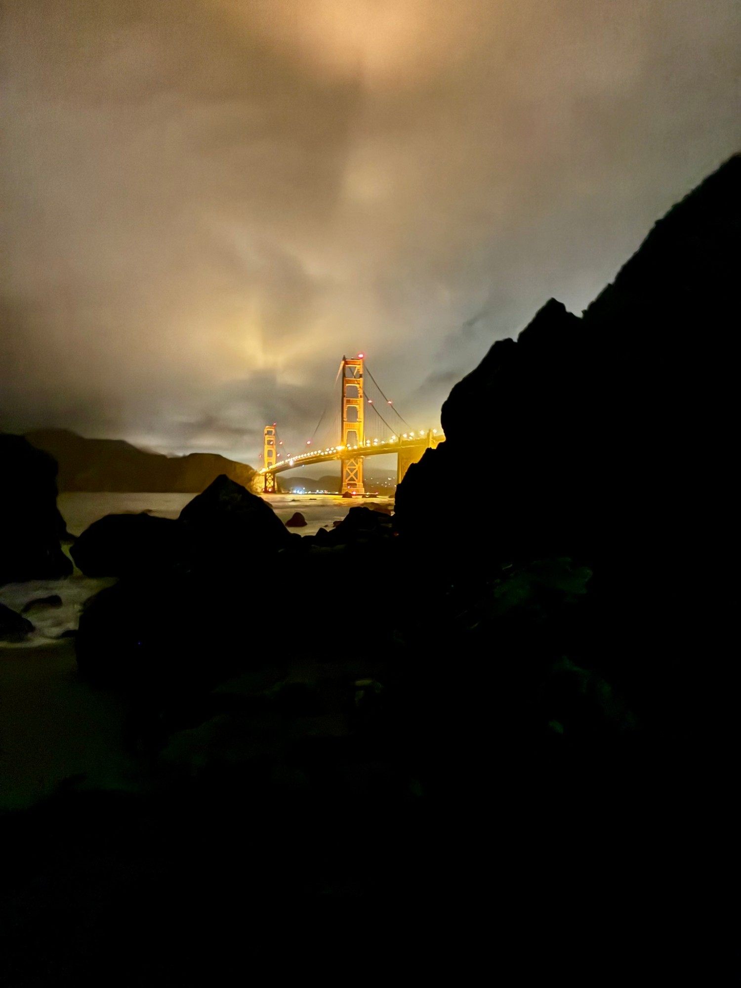 The illuminated Golden Gate Bridge seen between dark rock silhouettes on Marshall's Beach under glowing clouds