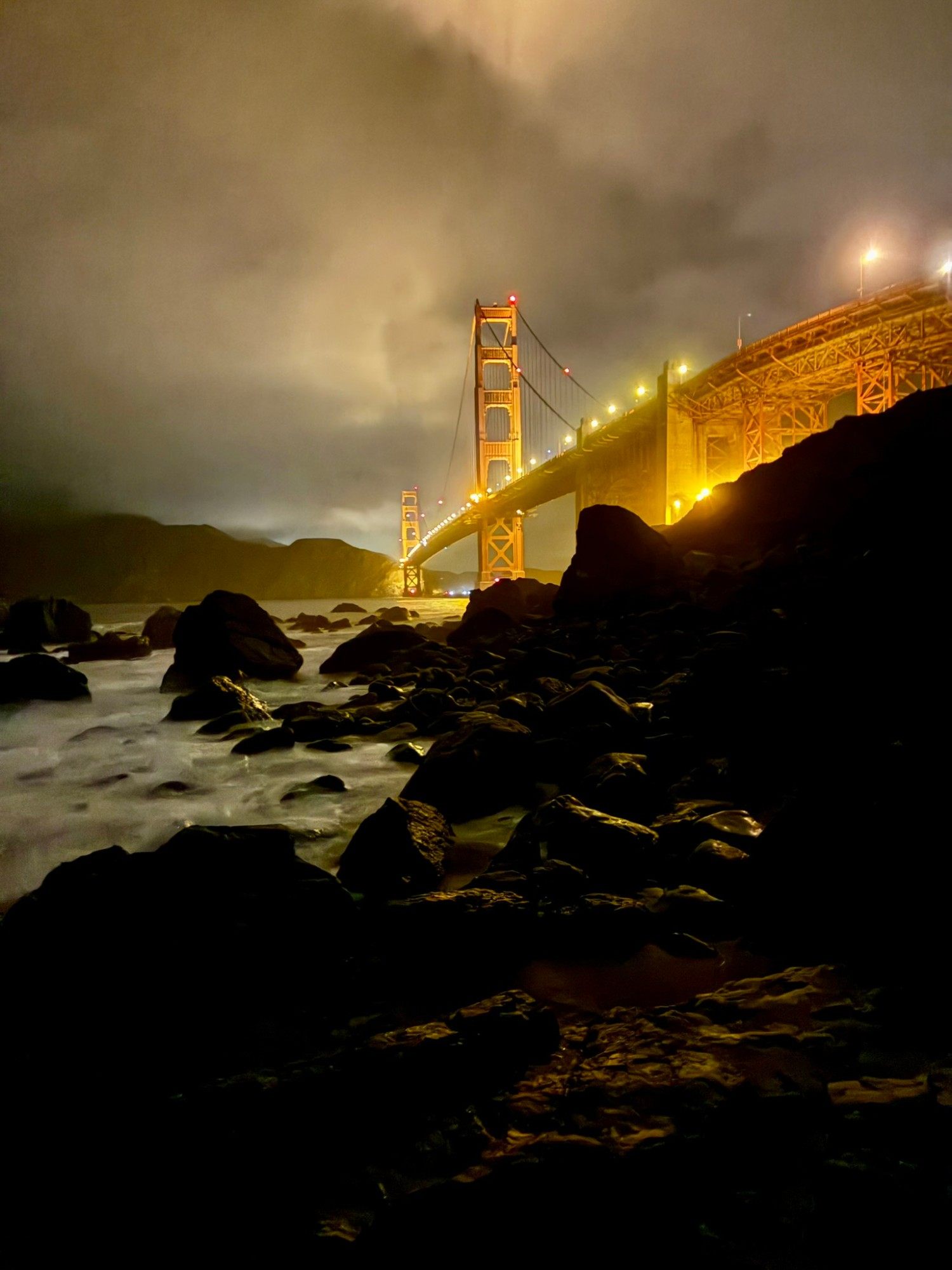 Wet boulders in the foreground and the Golden Gate Bridge lit up in gold above the rocky shoreline