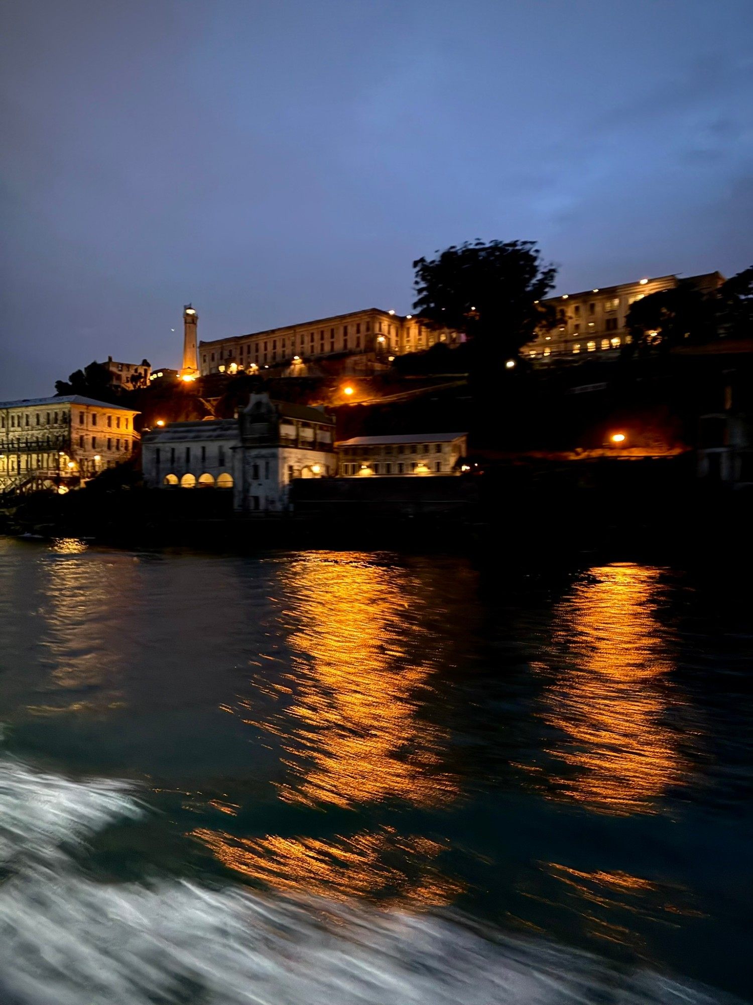 Alcatraz Island with its lighthouse and cellhouse lit up at dusk, with orange reflections in the water