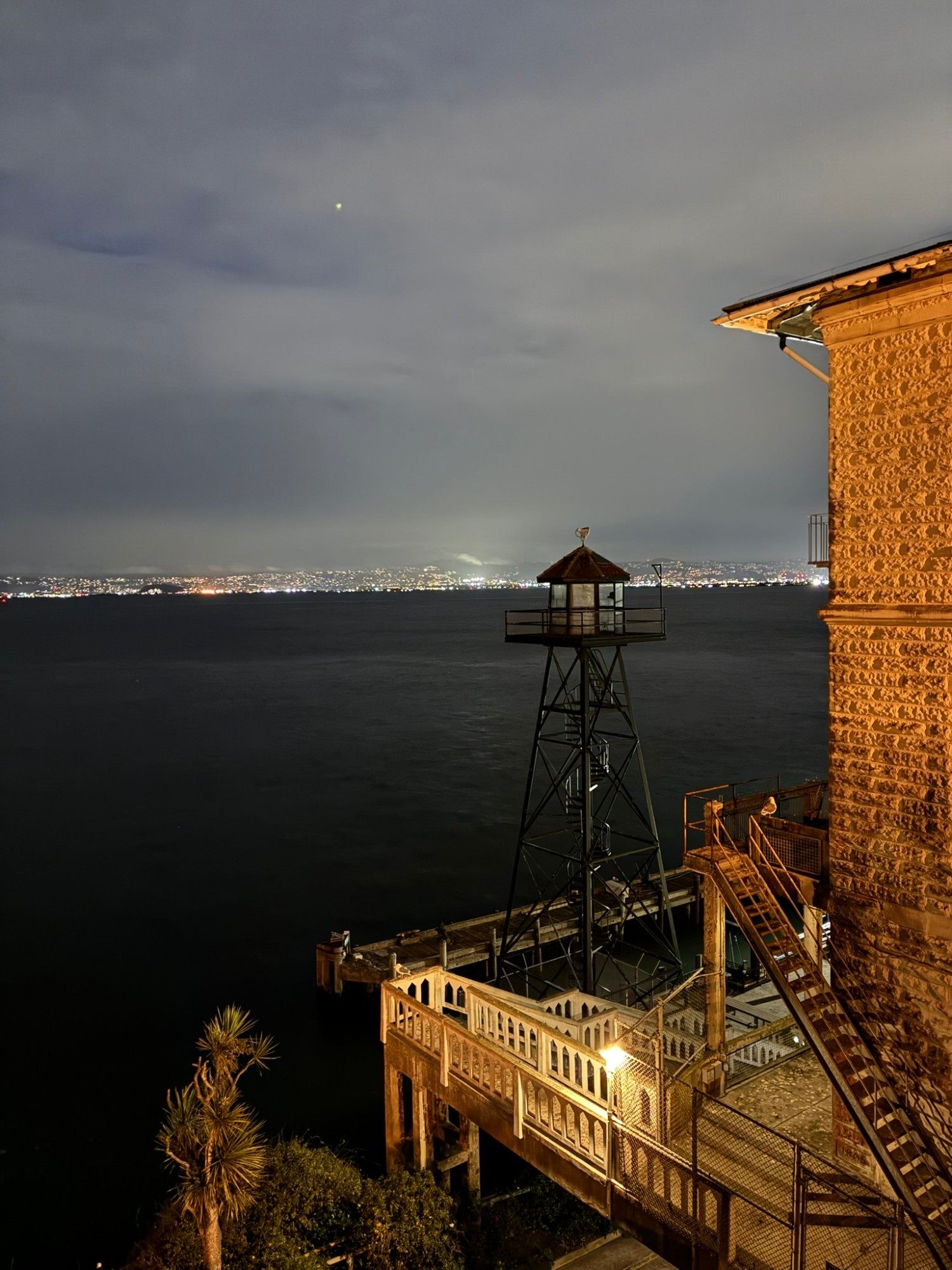 An old guard tower by the Alcatraz dock at night, with the lights of the bay shore across the dark water