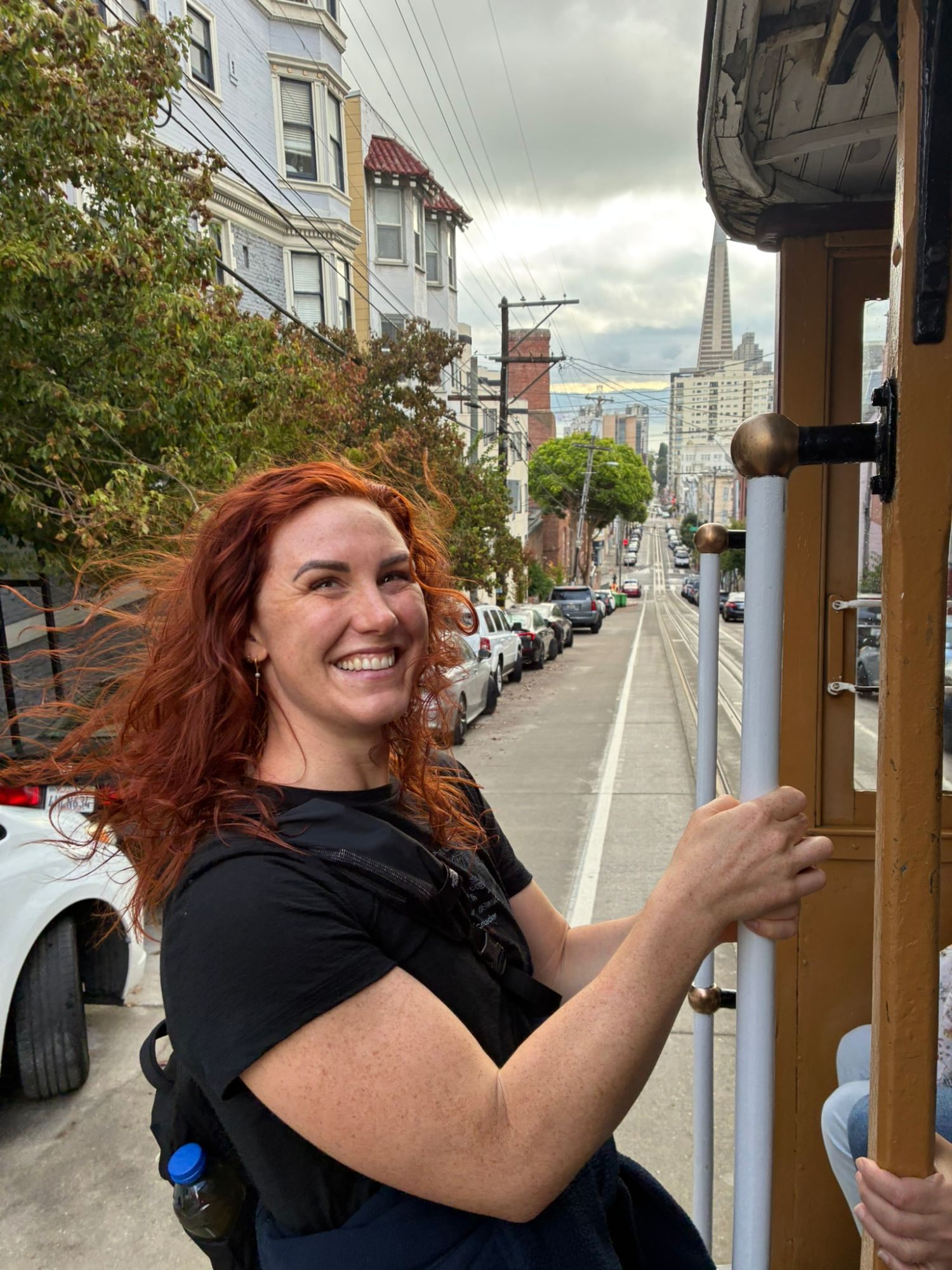 Kristine laughing as she holds on to the outside of a San Francisco cable car on a steep street, with the Transamerica Pyramid in the distance