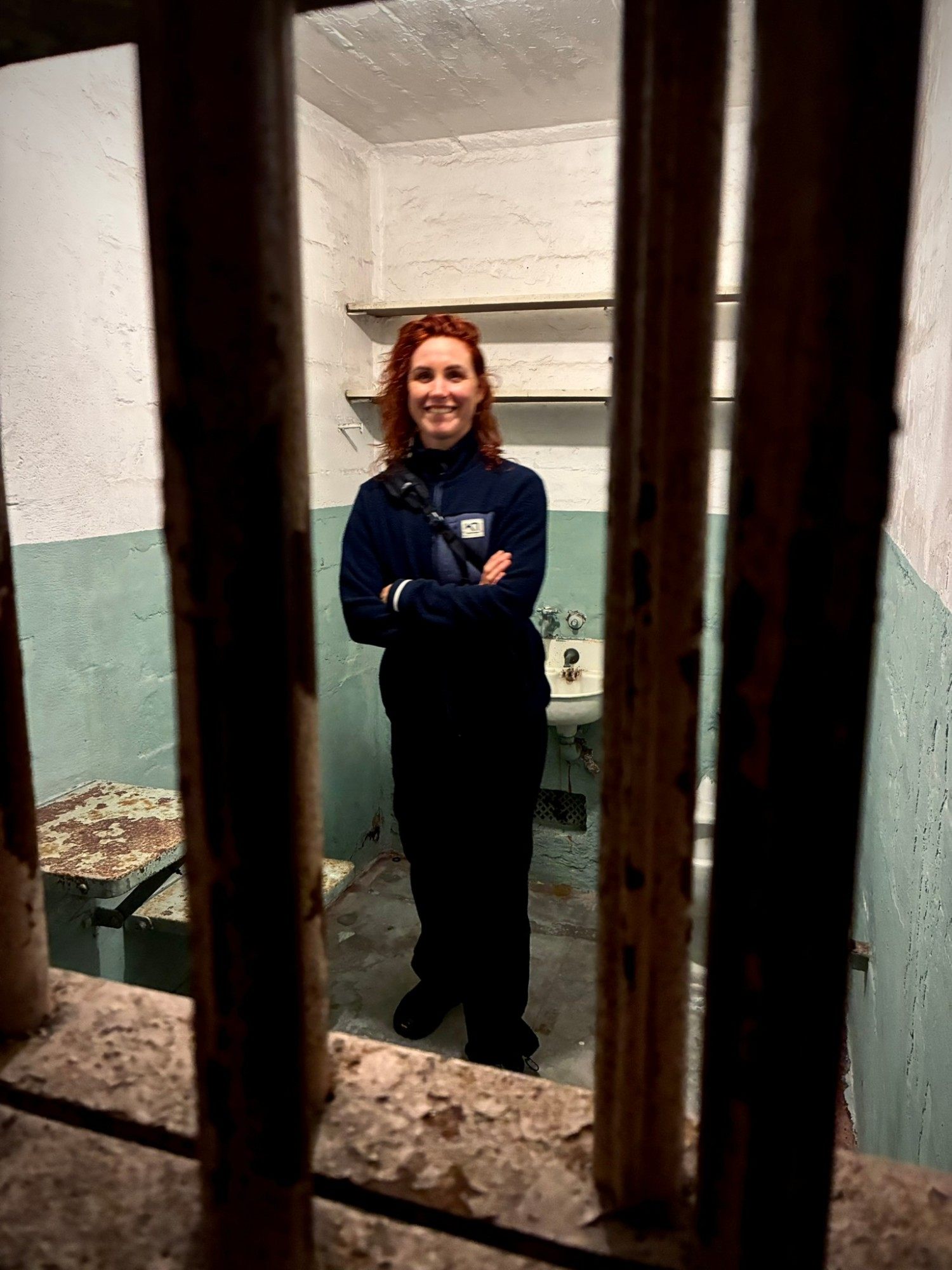 Kristine smiling with her arms crossed inside a small Alcatraz cell, photographed through the bars