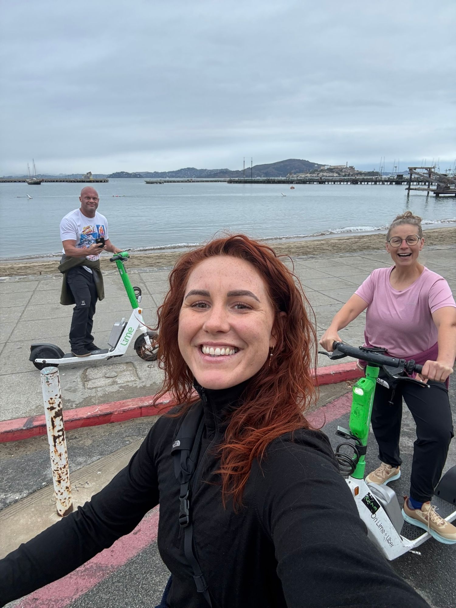 Selfie of Kristine with Inge and me on electric scooters by the water, with Alcatraz in the background