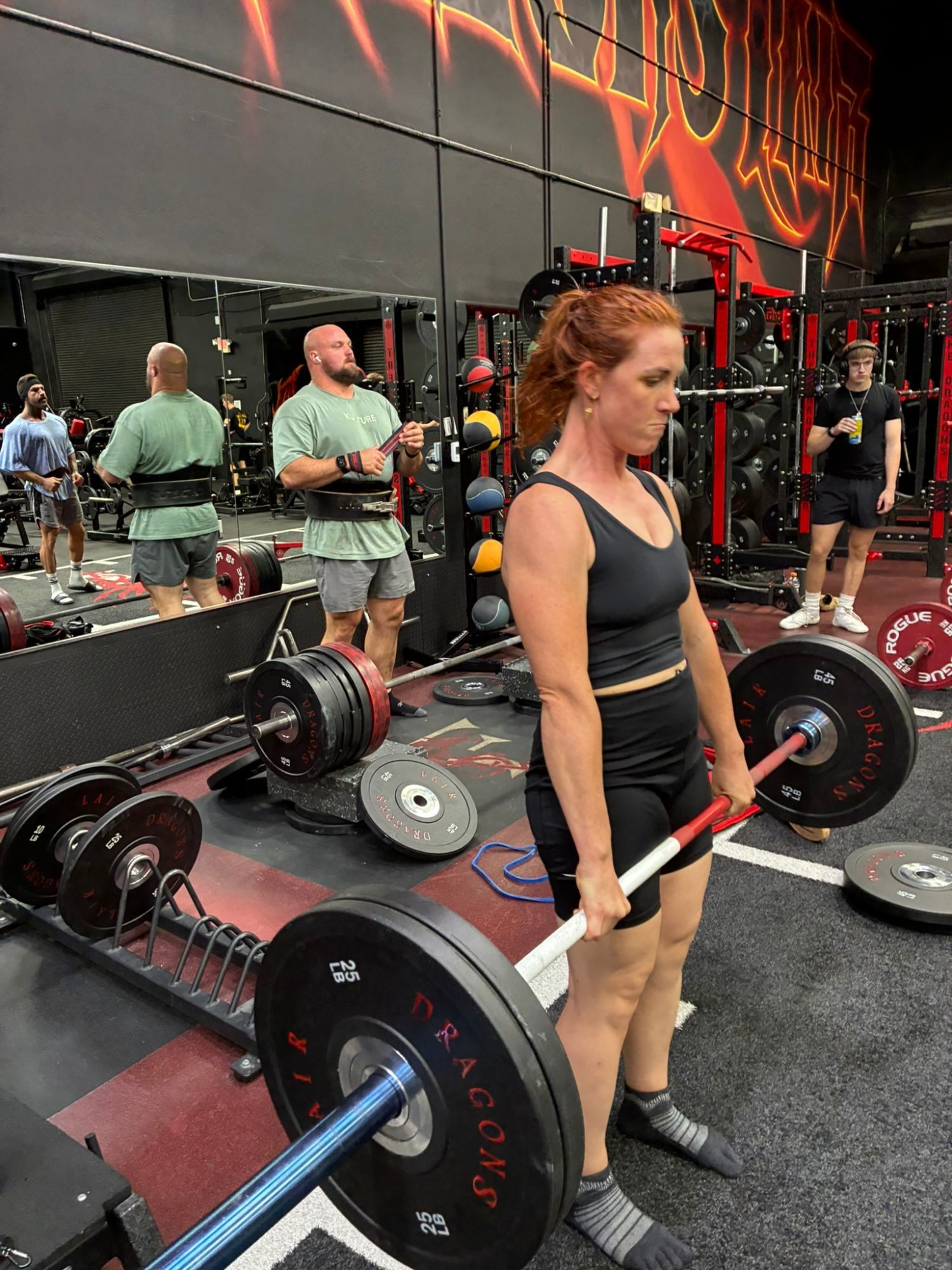 Kristine deadlifting a loaded barbell in socks, with Mitchell Hooper wrapping his wrists in the background