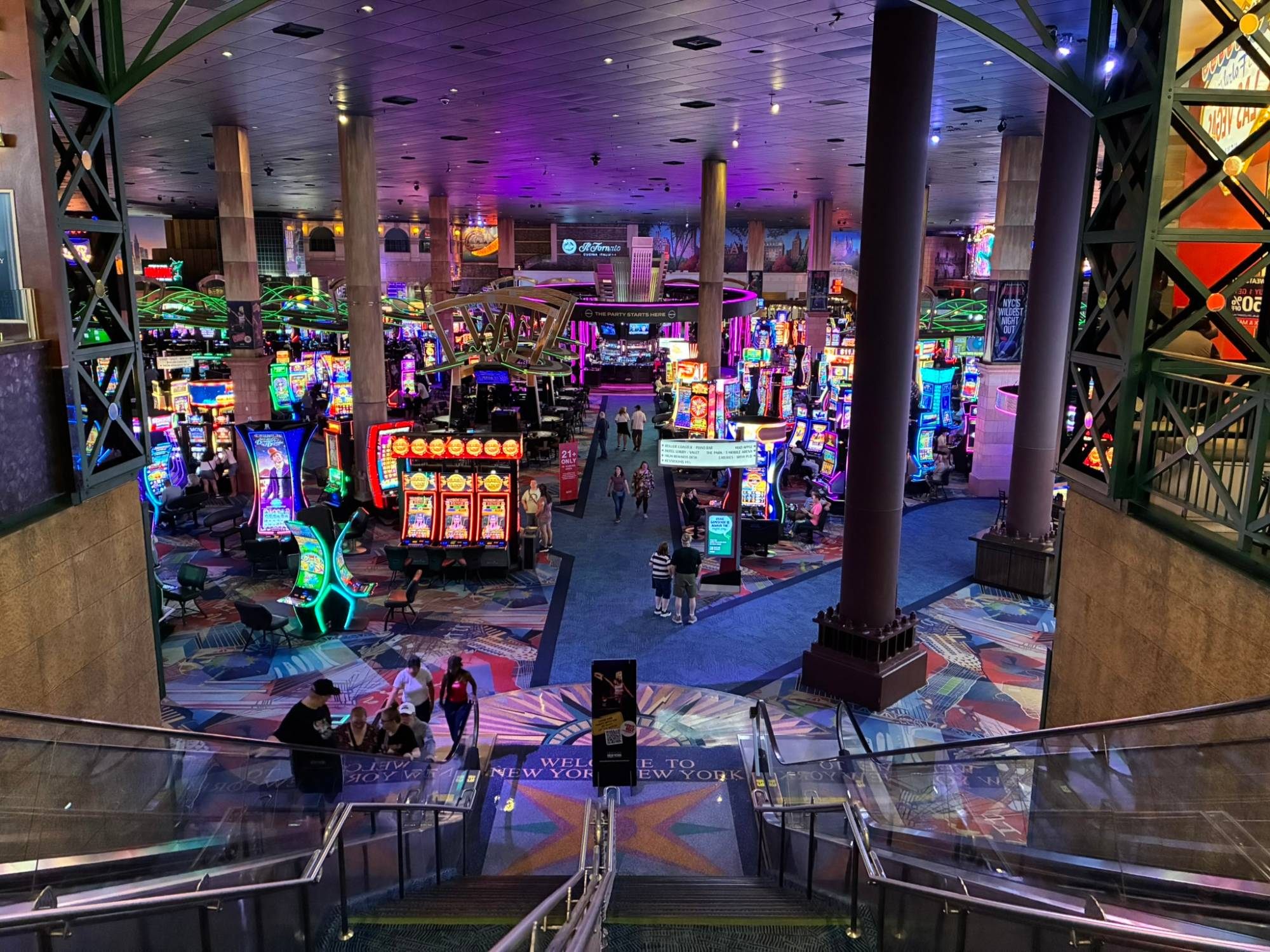 A huge casino floor seen from the top of an escalator, full of brightly lit slot machines under a purple ceiling, with a Welcome to New York New York mat at the bottom