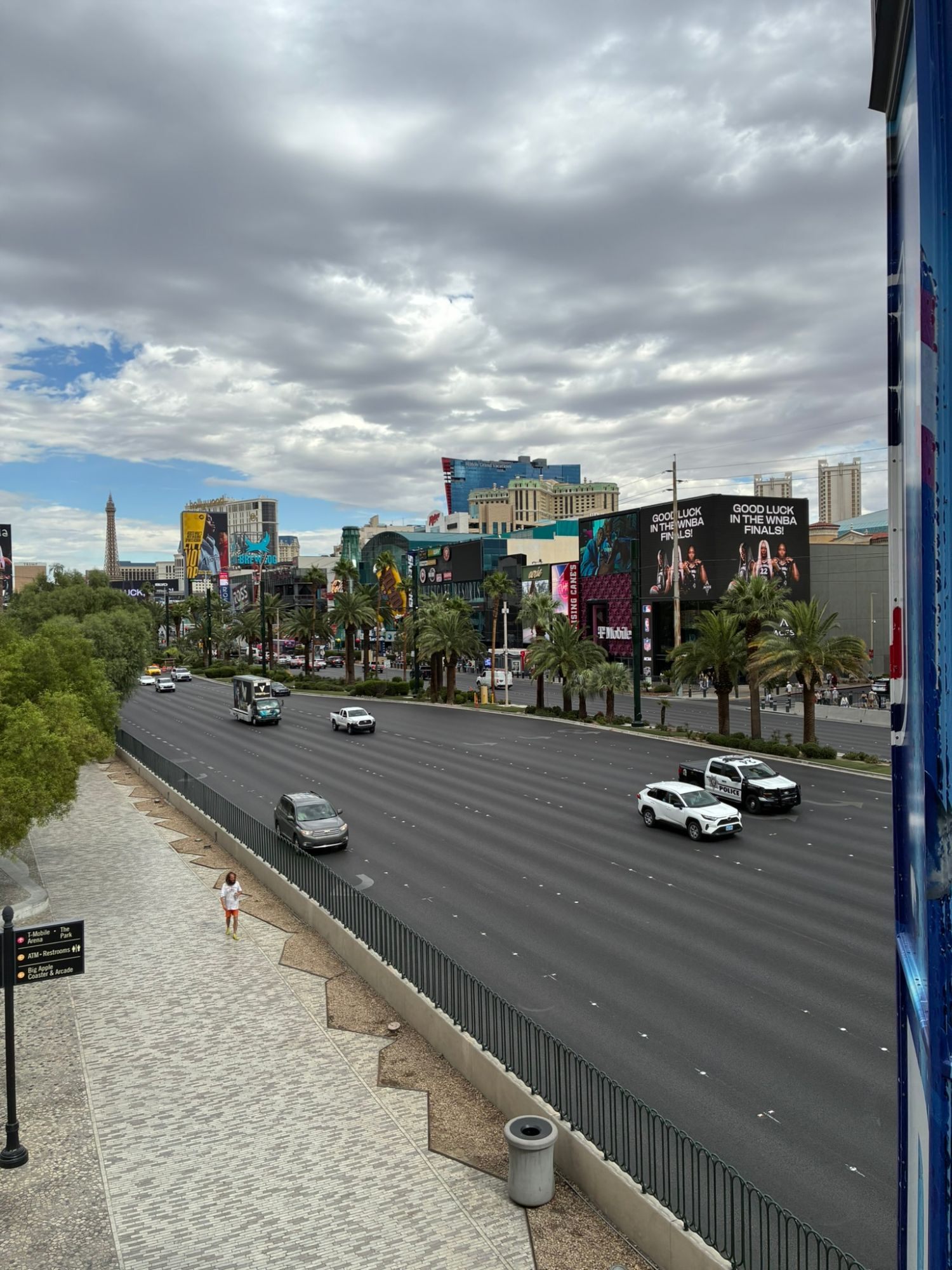 The wide multi-lane Las Vegas Strip seen from a pedestrian bridge, with palm trees, hotels, billboards and a replica Eiffel Tower in the distance